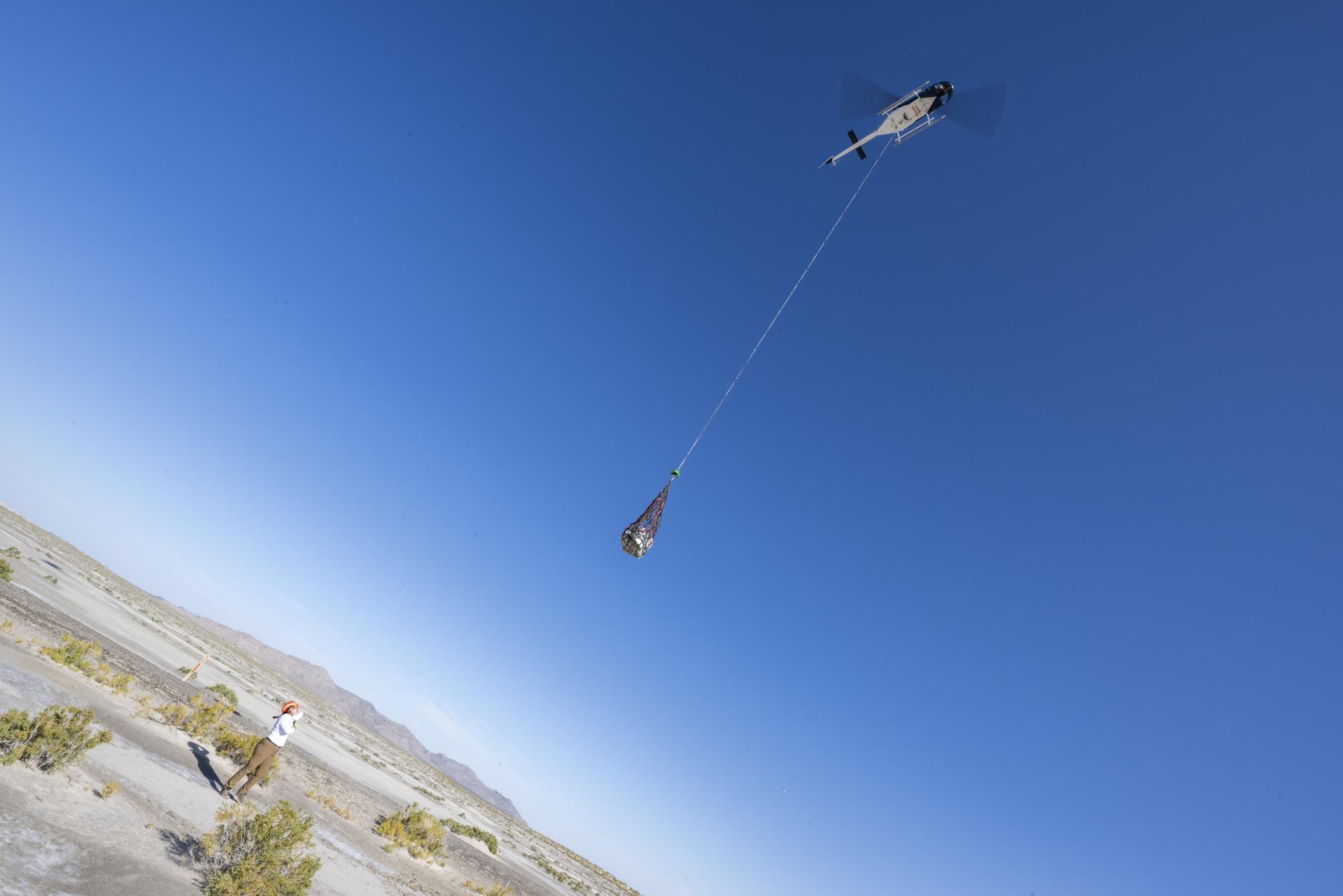 The OSIRIS-REx sample capsule hangs suspended from a helicopter by a long line against a bright blue sky and the desert below. A woman looks up at the capsule as it departs.