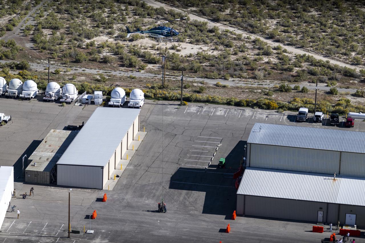 The sample return capsule from NASA’s OSIRIS-REx mission is lowered into the parking lot in front of the cleanroom hangar by helicopter, Sunday, Sept. 24, 2023, shortly after the capsule landed at the Department of Defense's Utah Test and Training Range. The sample was collected from the asteroid Bennu in October 2020 by NASA’s OSIRIS-REx spacecraft. Photo Credit: (NASA/Keegan Barber)