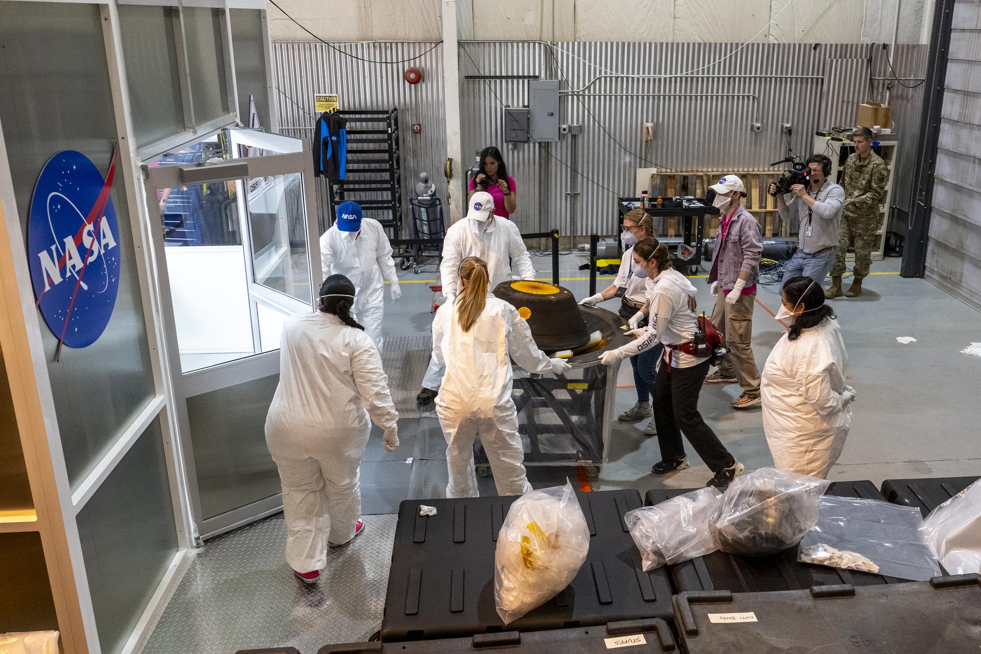 Several people, wearing various levels of protective gear, walk with a wheeled platform. The platform holds the OSIRIS-REx sample capsule, which is a squat black pentagon. They are wheeling the platform through a hangar toward an enclosed room with the NASA meatball logo.