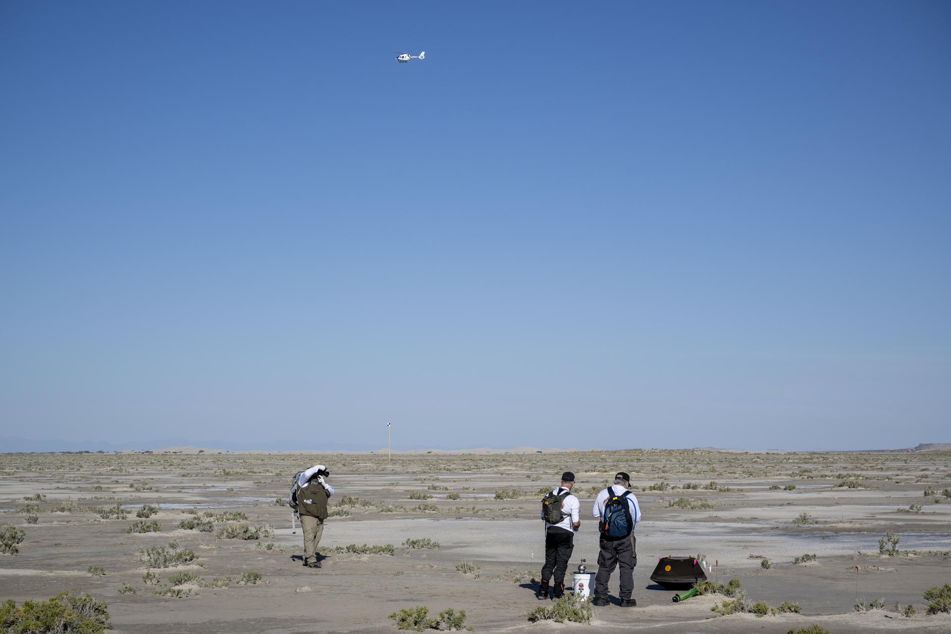Three people stand in the desert. A white helicopter flies away, against a blue sky.