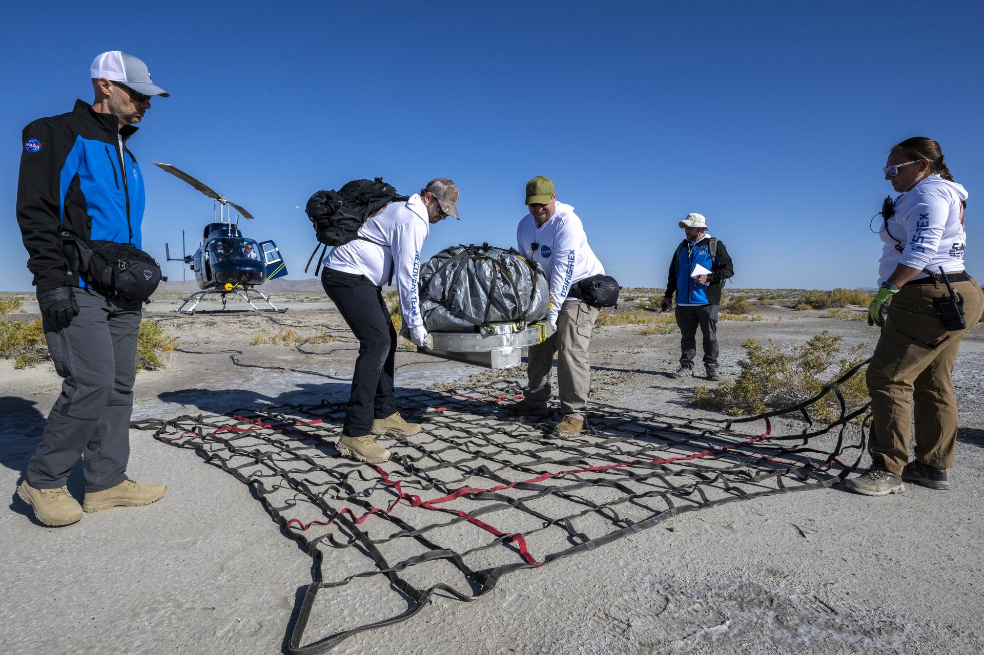 Two people carry the OSIRIS-REx sample capsule, now wrapped in protective tarp, onto a net laid on the desert ground. Three other people are standing nearby to help. A helicopter sits in the background.