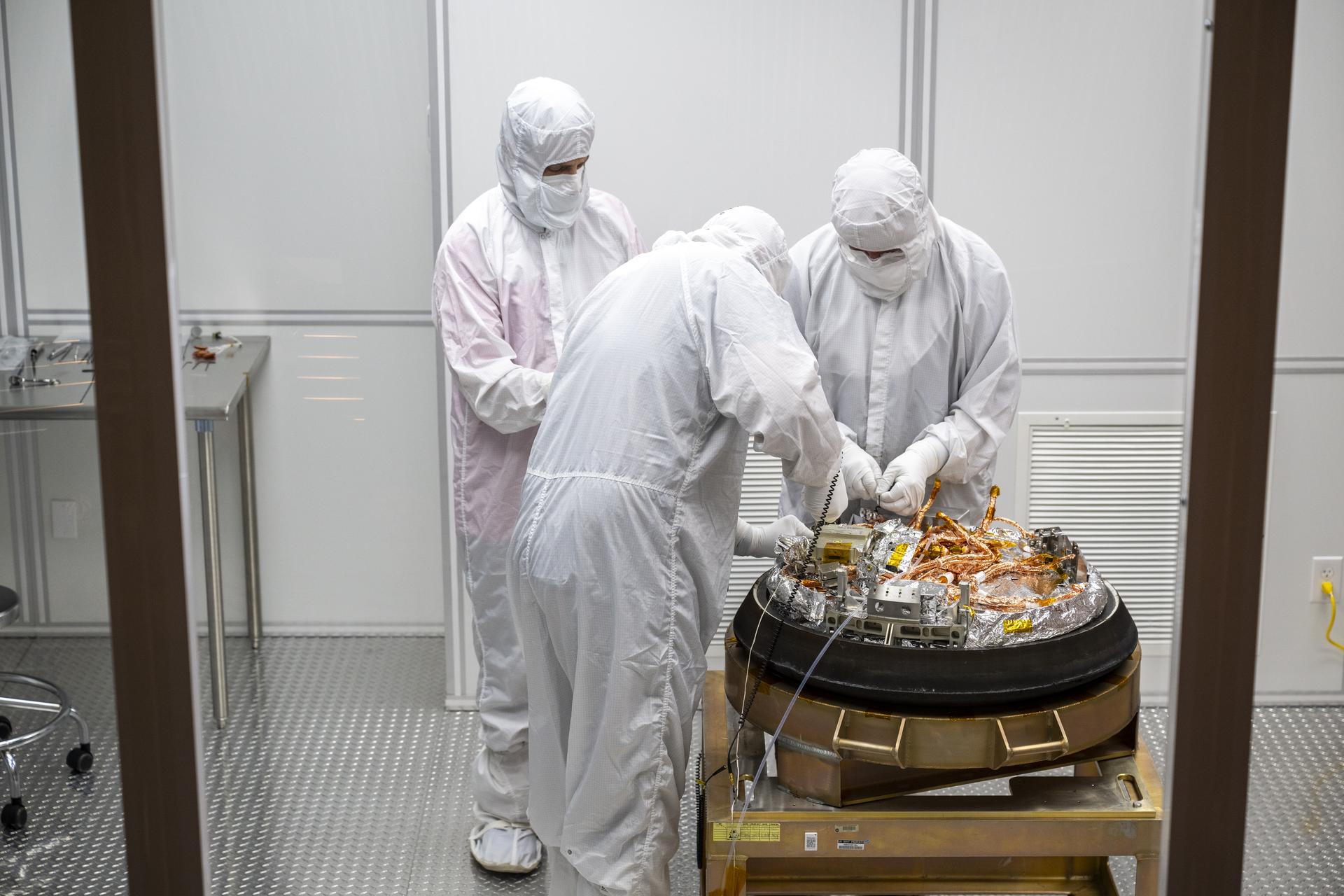 Three people in white clean room suits with hoods and masks inside a small, mobile clean room. They are working on the OSIRIS-REx sample capsule, which sits on a platform.