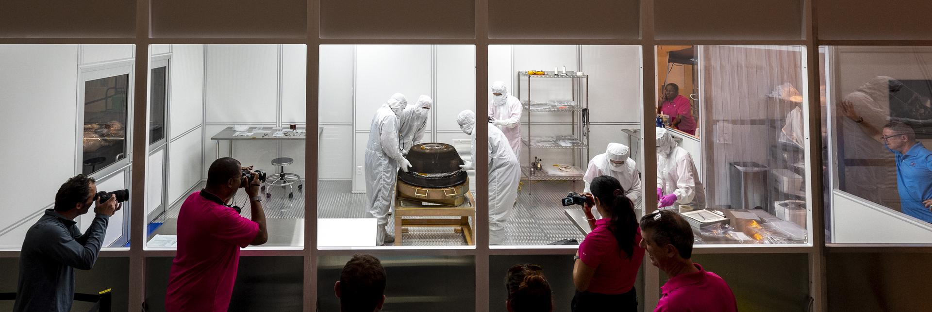 Several people stand outside a small, enclosed, windowed room, looking in. Inside the room, people in white clean room suits work on the OSIRIS-REx sample capsule, which sits on a platform.