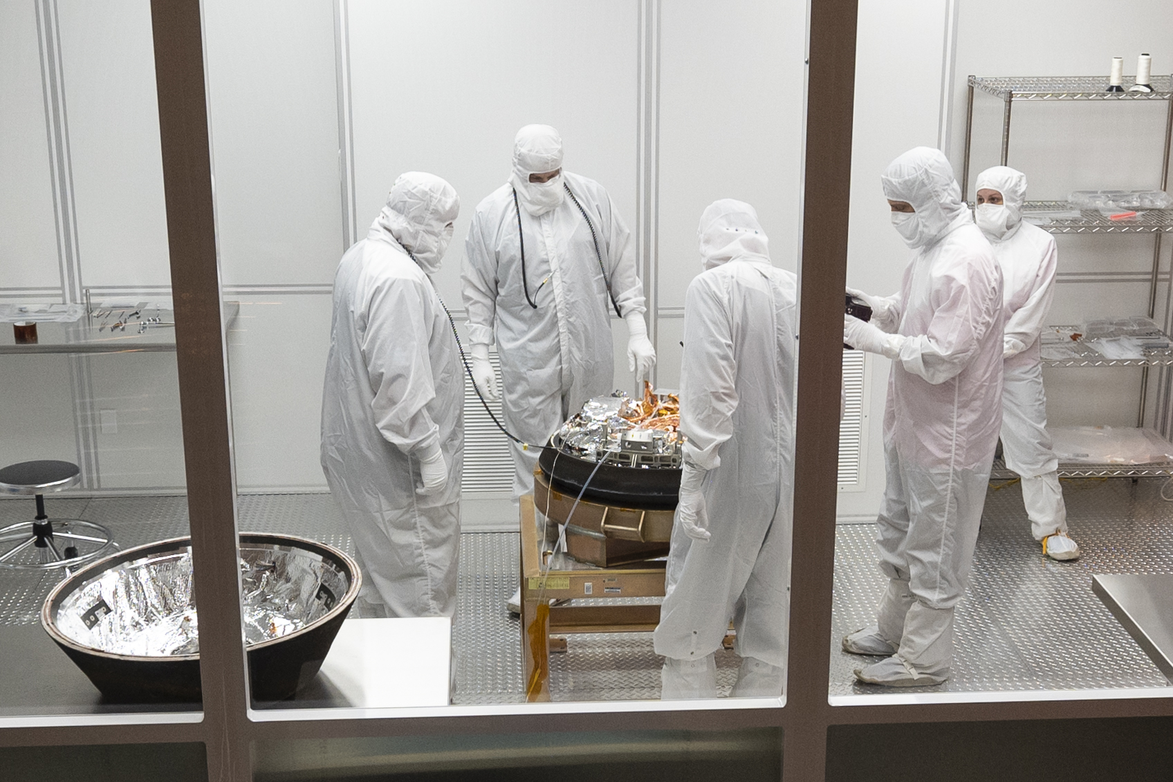 Five people in white clean room suits with hoods and masks inside a small, mobile clean room. They are working on the OSIRIS-REx sample capsule, which sits on a platform.
