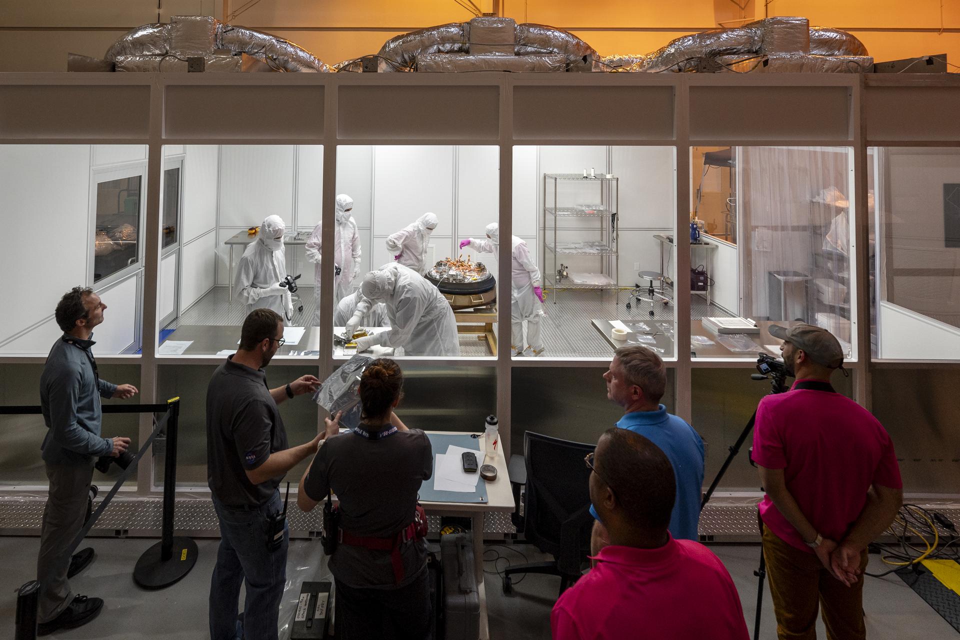 Several people stand outside a small, enclosed, windowed room, looking in. Inside the room, people in white clean room suits work on the OSIRIS-REx sample capsule, which sits on a platform.