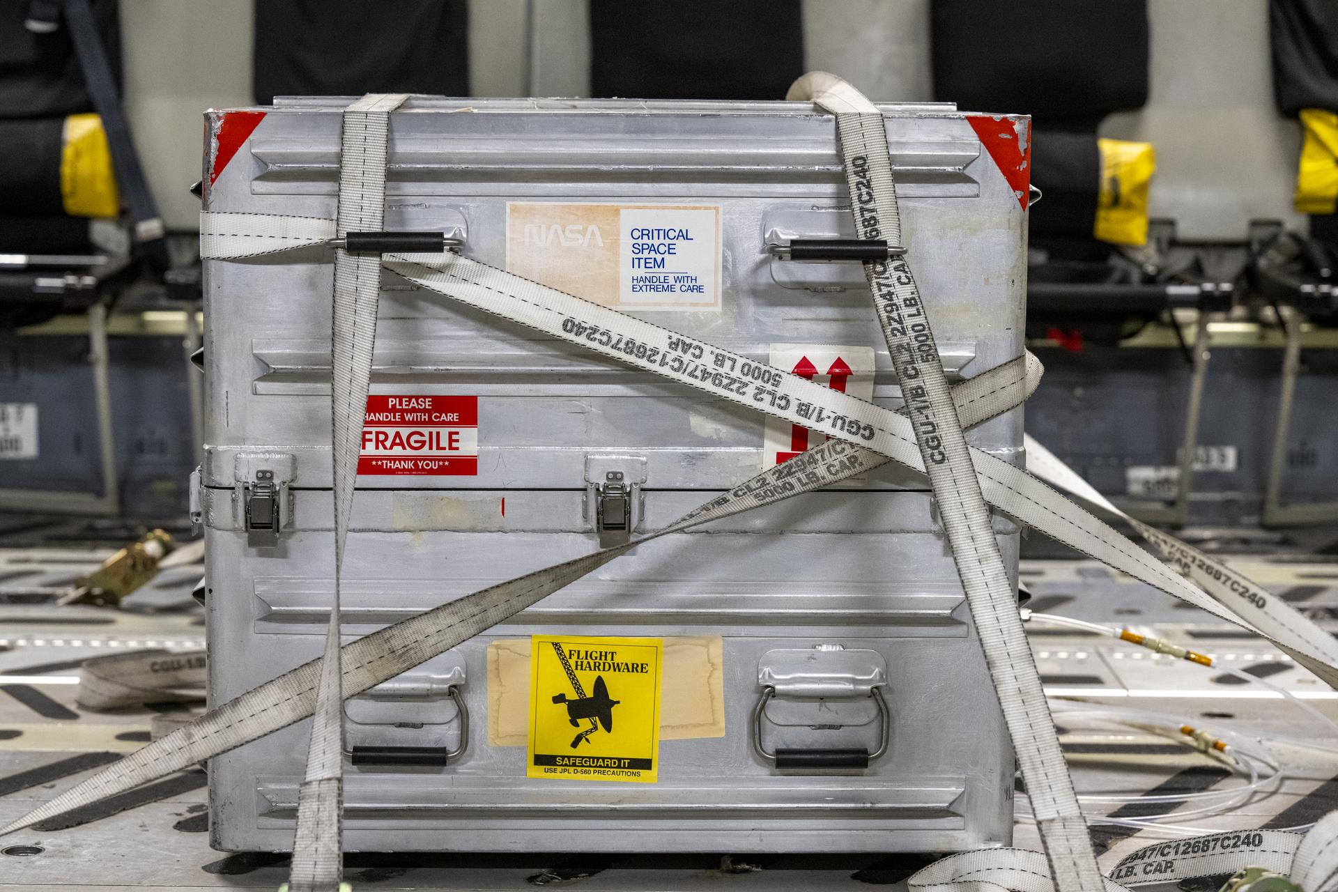 A silver box, strapped into place and decorated with stickers that indicate it is fragile and contains flight hardware.