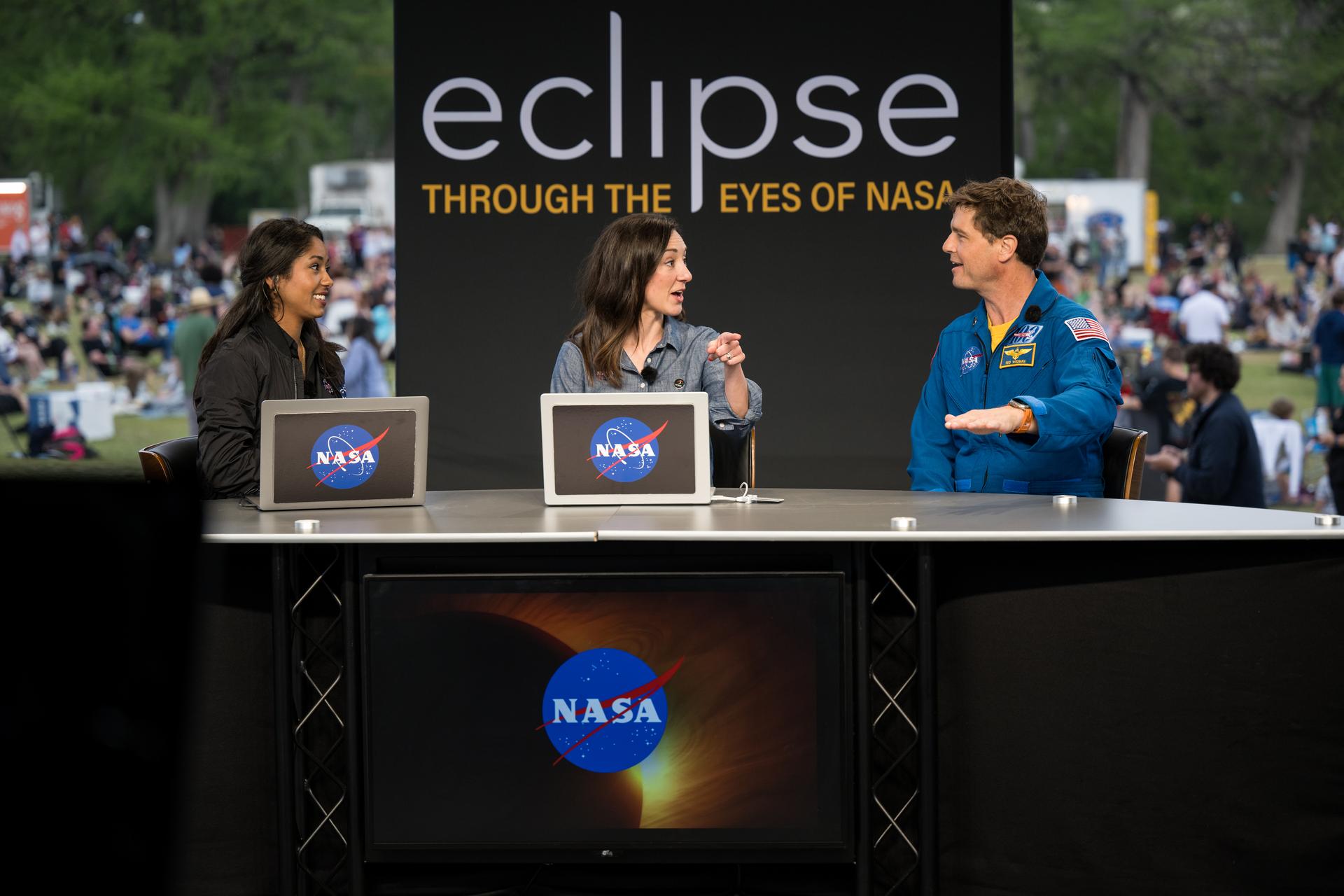 Three people sit at a news booth. One is wearing an astronaut jumpsuit.