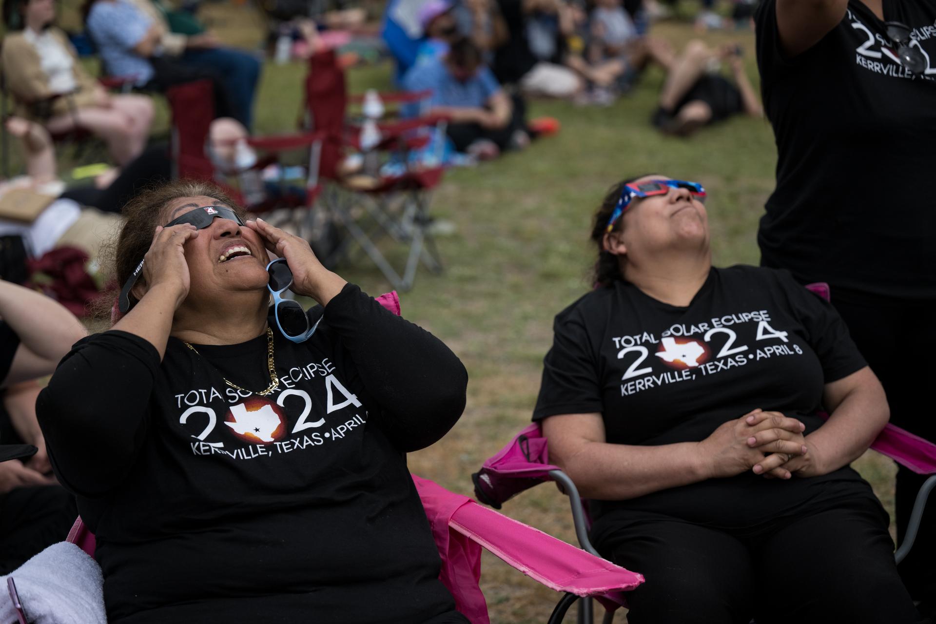 Two people wearing total solar eclipse shirts sit in chairs and look up at the sky while wearing eclipse glasses. Behind them are several other people sitting in chairs and on grass also watching the eclipse.