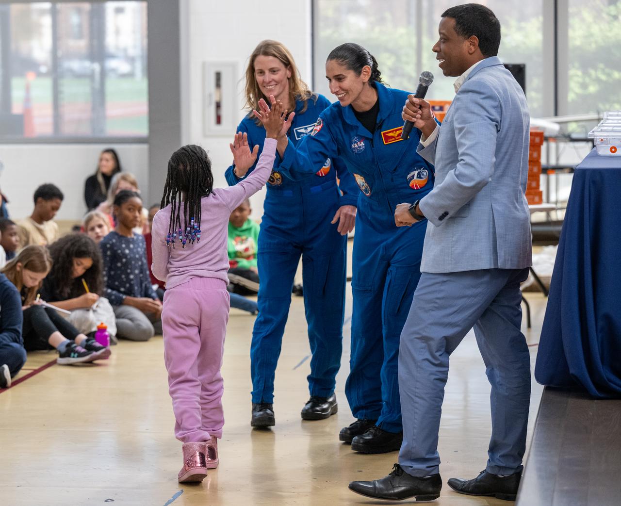 NASA astronauts Loral O’Hara and Jasmin Moghbeli, seen in blue flight suits, high five a student during an engagement event at Catherine Watkins Elementary School Monday, Dec. 2, 2024 in Washington. O’Hara and Moghbeli spent six months in space as part of Expedition 70 aboard the International Space Station. Photo Credit: (NASA/Keegan Barber)