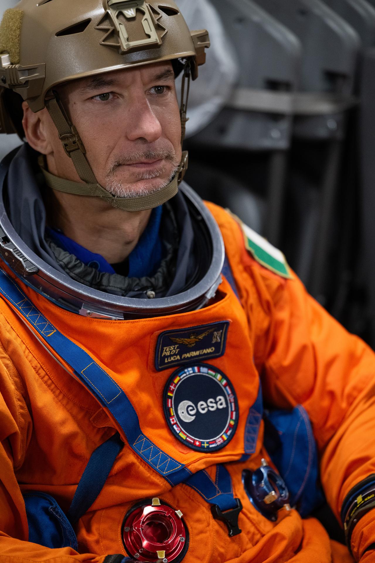A portrait of astronaut Luca Parmitano. He is wearing an orange space suit and a dull green helmet. Luca looks off-camera with a focused expression.