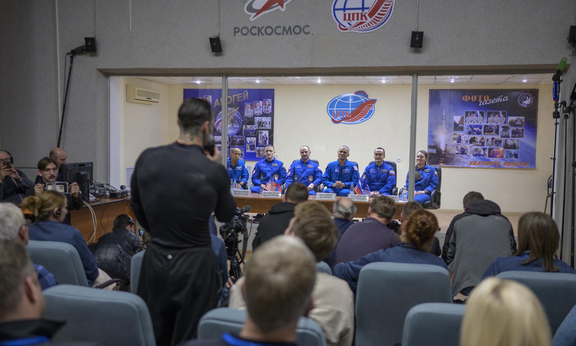 NASA astronaut Chris Williams, left, and Roscosmos cosmonauts Sergey Kud-Sverchkov, and Sergei Mikaev, along with Expedition 74 backup crew members Anil Menon of NASA, Roscosmos cosmonauts Petr Dubrov, and Anna Kikinam, right, are seen in quarantine, behind glass, as they arrive at a press conference, Wednesday, Nov. 26, 2025 at the Cosmonaut Hotel in Baikonur, Kazakhstan. Expedition 74 crewmembers: NASA astronaut Chris Williams, Roscosmos cosmonauts Sergey Kud-Sverchkov and Sergei Mikaev, are scheduled to launch aboard their Soyuz MS-28 spacecraft on November 27. Photo Credit: (NASA/Bill Ingalls)