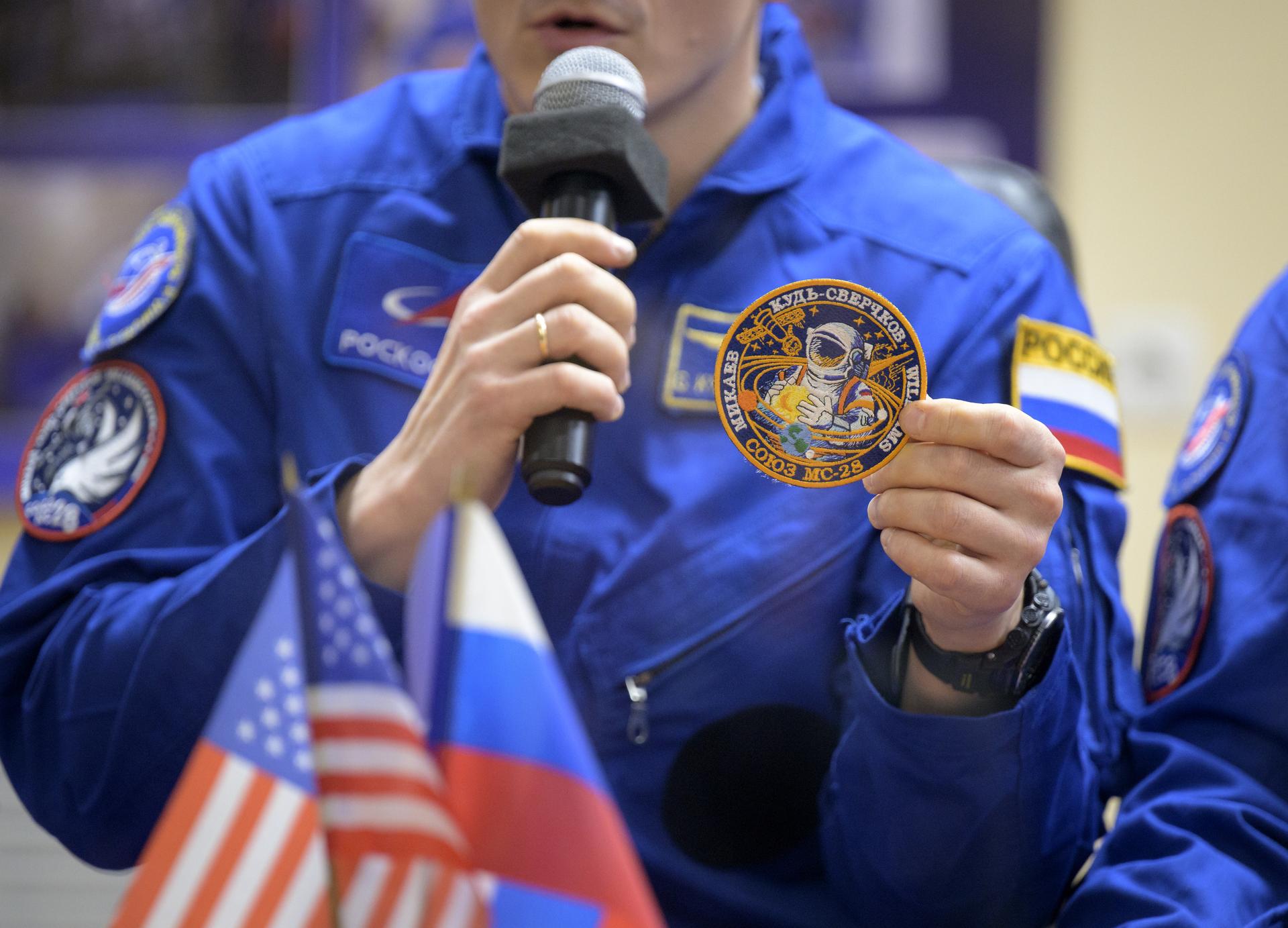 Roscosmos cosmonaut Sergey Kud-Sverchkov discusses his crew’s Soyuz patch while in quarantine, behind glass, during a press conference, Wednesday, Nov. 26, 2025 at the Cosmonaut Hotel in Baikonur, Kazakhstan. Expedition 74 crewmembers: NASA astronaut Chris Williams, Roscosmos cosmonauts Sergey Kud-Sverchkov and Sergei Mikaev, are scheduled to launch aboard their Soyuz MS-28 spacecraft on November 27. Photo Credit: (NASA/Bill Ingalls)