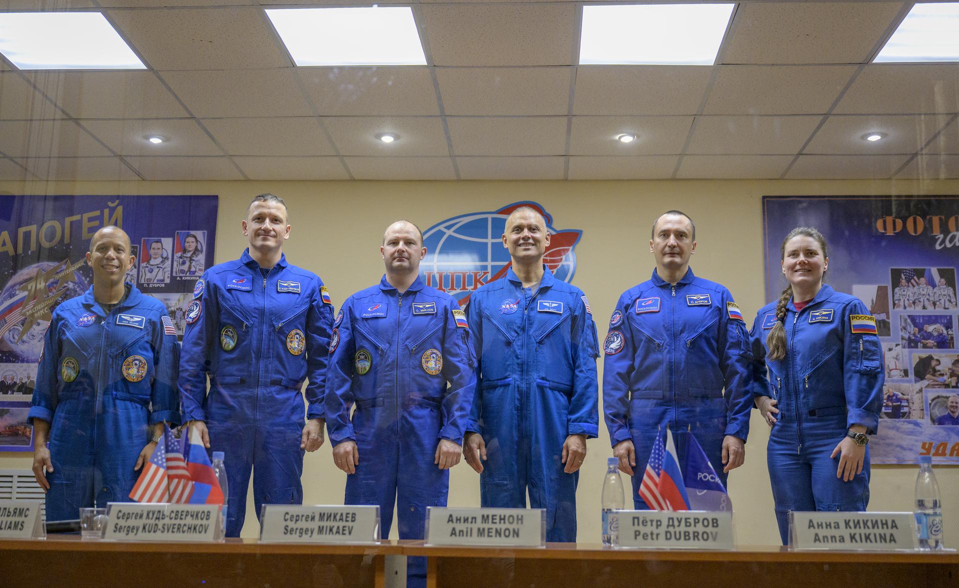 Expedition 74 price crew members: NASA astronaut Chris Williams, left, and Roscosmos cosmonauts Sergey Kud-Sverchkov, and Sergei Mikaev, along with Expedition 74 backup crew members Anil Menon of NASA, Roscosmos cosmonauts Petr Dubrov, and Anna Kikinam, right, are seen in quarantine, behind glass, as they finish a press conference, Wednesday, Nov. 26, 2025 at the Cosmonaut Hotel in Baikonur, Kazakhstan. Expedition 74 crewmembers: NASA astronaut Chris Williams, Roscosmos cosmonauts Sergey Kud-Sverchkov and Sergei Mikaev, are scheduled to launch aboard their Soyuz MS-28 spacecraft on November 27. Photo Credit: (NASA/Bill Ingalls)