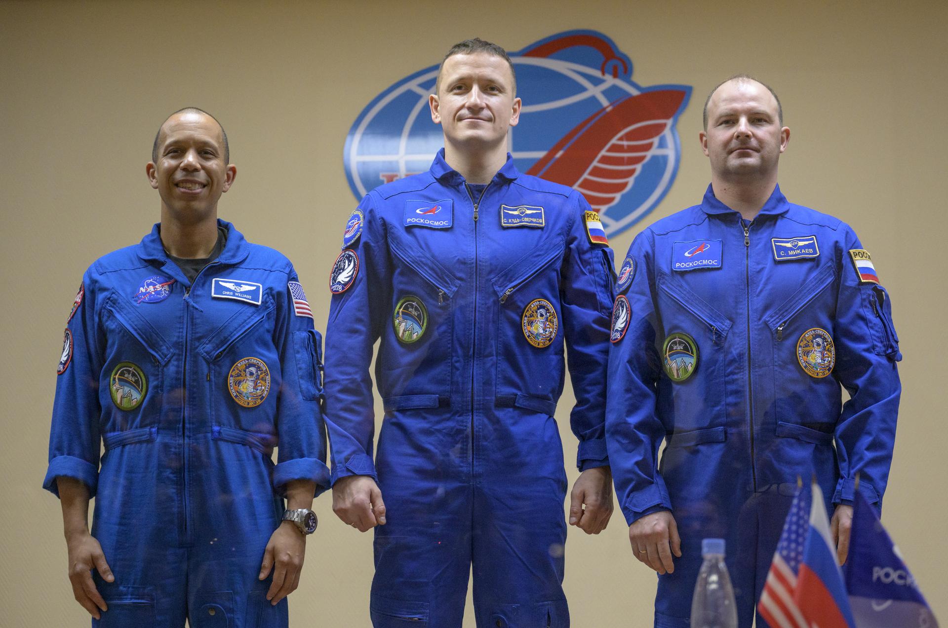 NASA astronaut Chris Williams, left, and Roscosmos cosmonauts Sergey Kud-Sverchkov, and Sergei Mikaev, right, are seen in quarantine, behind glass, at the conclusion of a press conference, Wednesday, Nov. 26, 2025 at the Cosmonaut Hotel in Baikonur, Kazakhstan. Expedition 74 crewmembers: NASA astronaut Chris Williams, Roscosmos cosmonauts Sergey Kud-Sverchkov and Sergei Mikaev, are scheduled to launch aboard their Soyuz MS-28 spacecraft on November 27. Photo Credit: (NASA/Bill Ingalls)