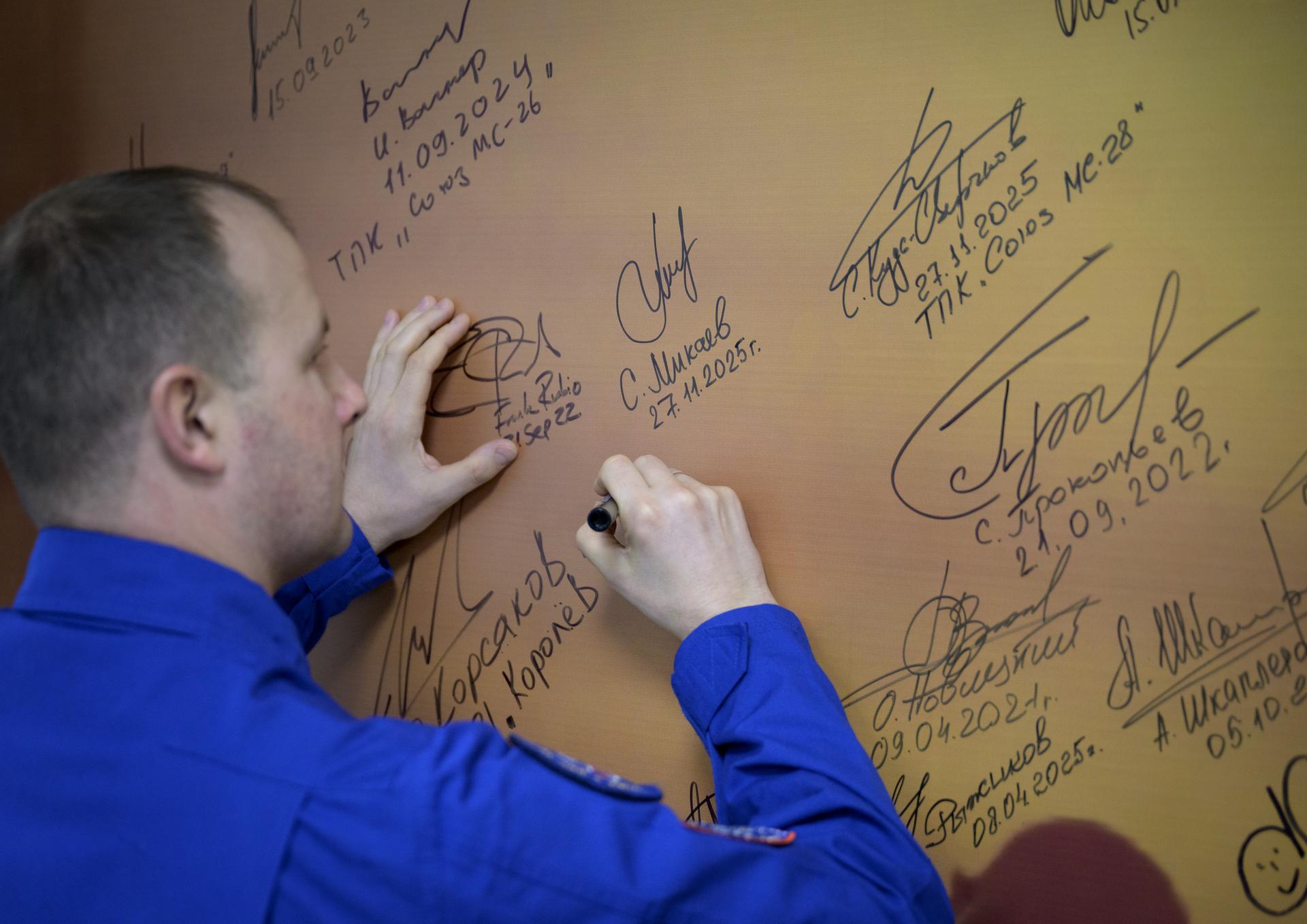 Roscosmos cosmonaut Sergei Mikaev signs a mural at the Cosmonaut Hotel prior to departing for suit-up and launch on a Soyuz rocket with fellow crewmates NASA astronaut Chris Williams, and Roscosmos cosmonaut Sergey Kud-Sverchkov, Thursday, Nov. 27, 2025, in Baikonur, Kazakhstan. The launch will send Williams, Kud-Sverchkov and Mikaev, on a mission to the International Space Station. Photo Credit: (NASA/Bill Ingalls)
