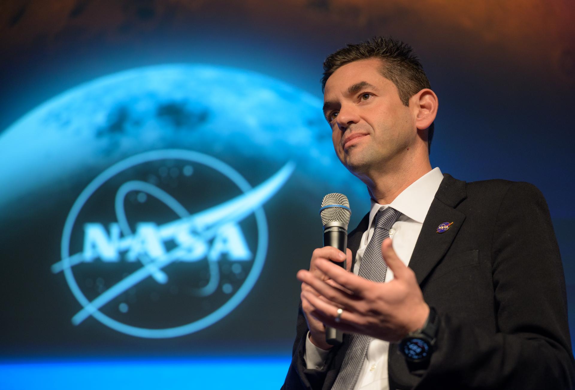 NASA Administrator Jared Isaacman smiling in a dark suit and blue tie, standing in front of an American flag and NASA logo backdrop.