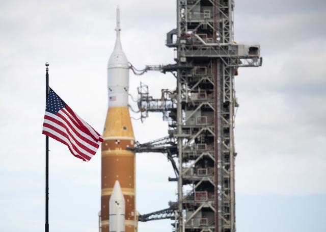 An American flag is seen in the foreground as NASA’s Space Launch System (SLS...