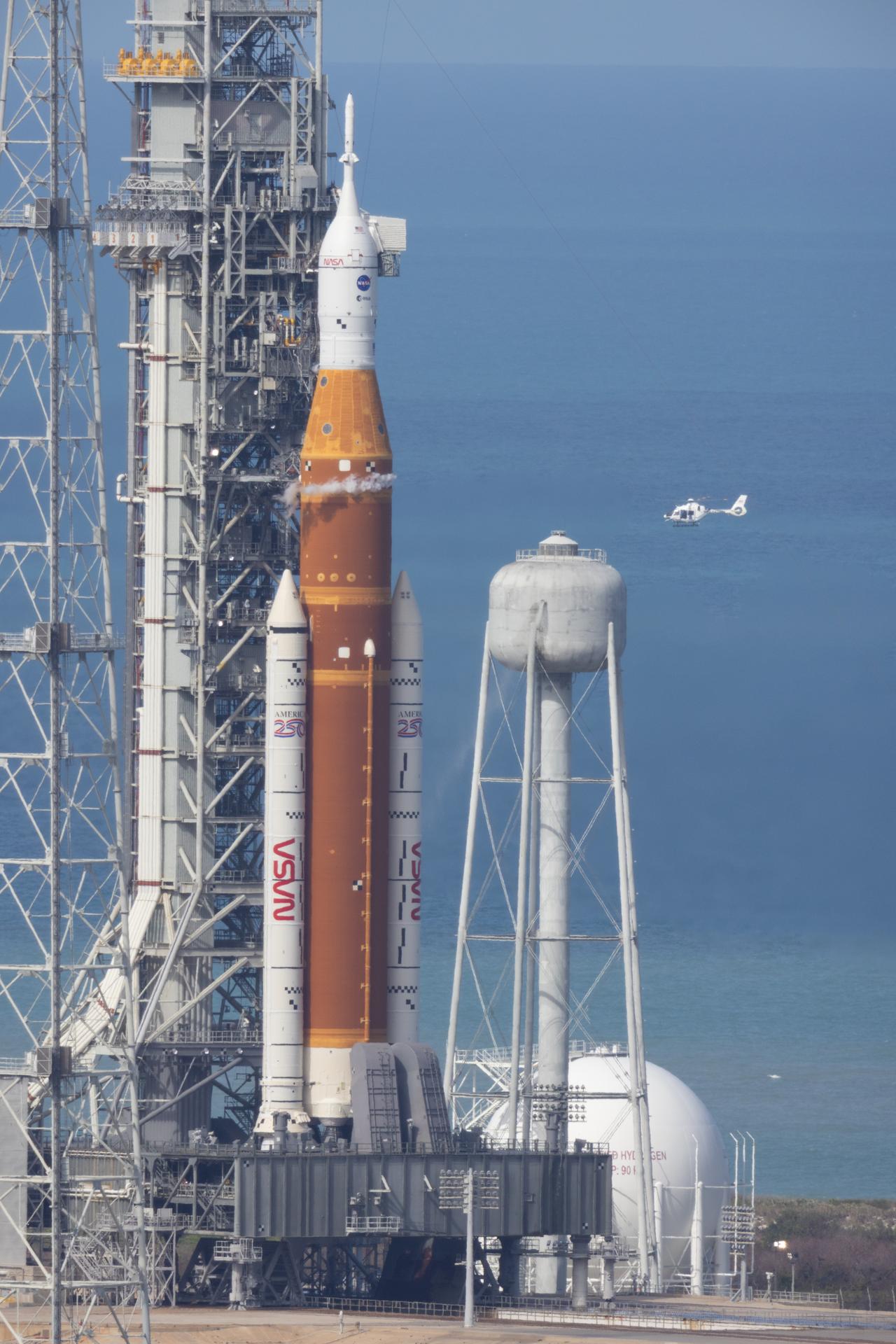 A NASA helicopter flies past the agency’s Space Launch System (SLS) rocket with the Orion spacecraft aboard is seen atop the mobile launcher at Launch Complex 39B, Wednesday, April 1, 2026, as the launch countdown progresses at NASA’s Kennedy Space Center in Florida. NASA’s Artemis II mission will take NASA astronauts Reid Wiseman, commander; Victor Glover, pilot; Christina Koch, mission specialist; and CSA (Canadian Space Agency) astronaut Jeremy Hansen, mission specialist on a 10-day journey around the Moon and back aboard SLS rocket and Orion spacecraft from Launch Complex 39B, with a two hour launch window opening at 6:24 p.m. EDT.  Photo Credit: (NASA/Joel Kowsky)
