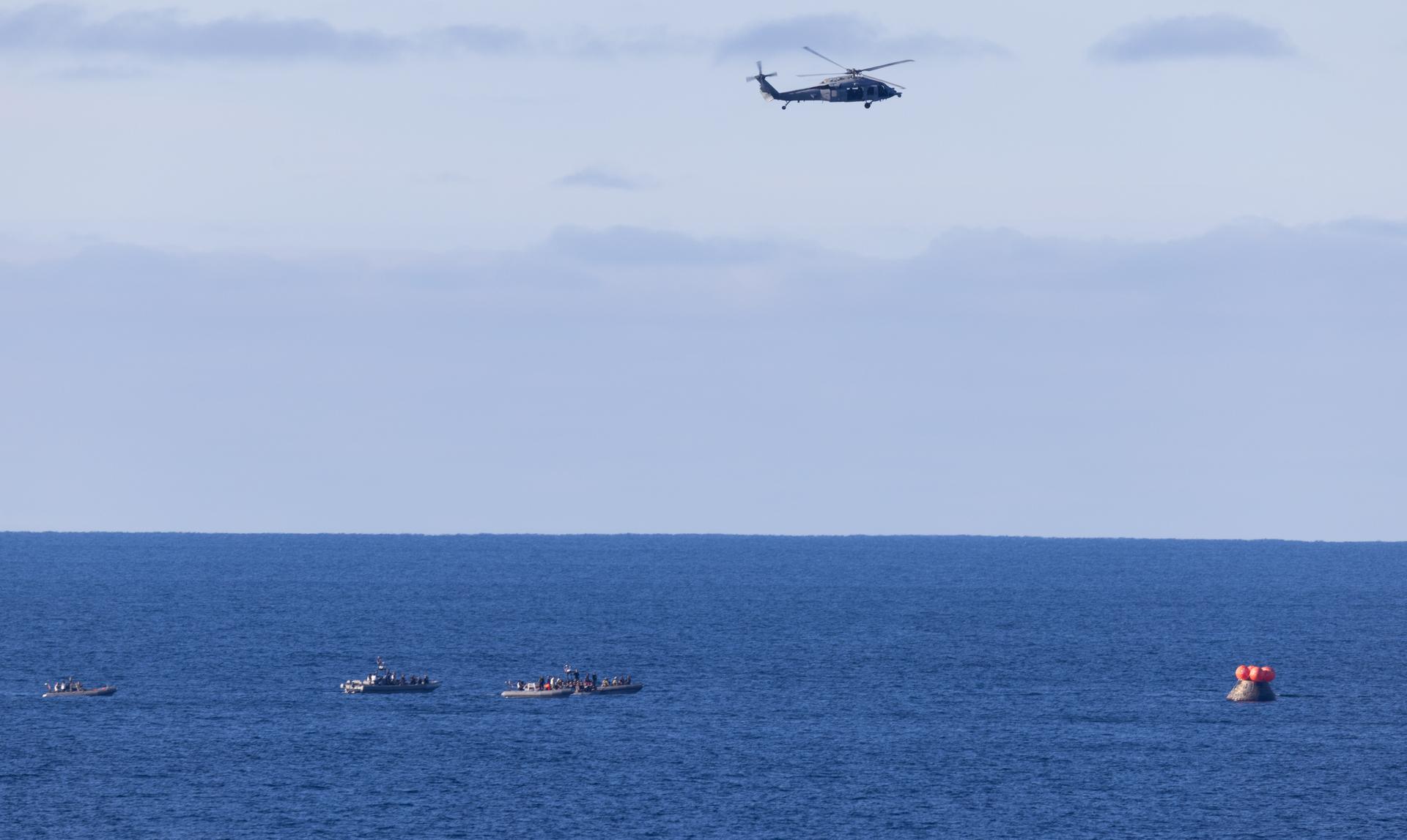 A U.S. Navy MH-60 Seahawks from Helicopter Sea Combat Squadron (HSC) 23 flies overhead as small boats approach NASA’s Orion spacecraft with Artemis II crewmembers NASA astronauts Reid Wiseman, commander; Victor Glover, pilot; Christina Koch, mission specialist; and CSA (Canadian Space Agency) astronaut Jeremy Hansen, mission specialist aboard is seen after landing as recovery teams as NASA’s Landing and Recovery team, along with U.S. Navy personnel in small boats begin to approach the spacecraft in the Pacific Ocean off the coast of California, Friday, April 10, 2026. NASA’s Artemis II mission took Wiseman, Glover, Koch, and Hansen on a nearly 10-day journey around the Moon and back to Earth. Following a splashdown at 5:07 p.m. PDT (8:07 p.m. EDT), NASA and U.S. military teams are working to bring the crewmembers and Orion spacecraft aboard USS John P. Murtha.  Photo Credit: (NASA/Joel Kowsky)