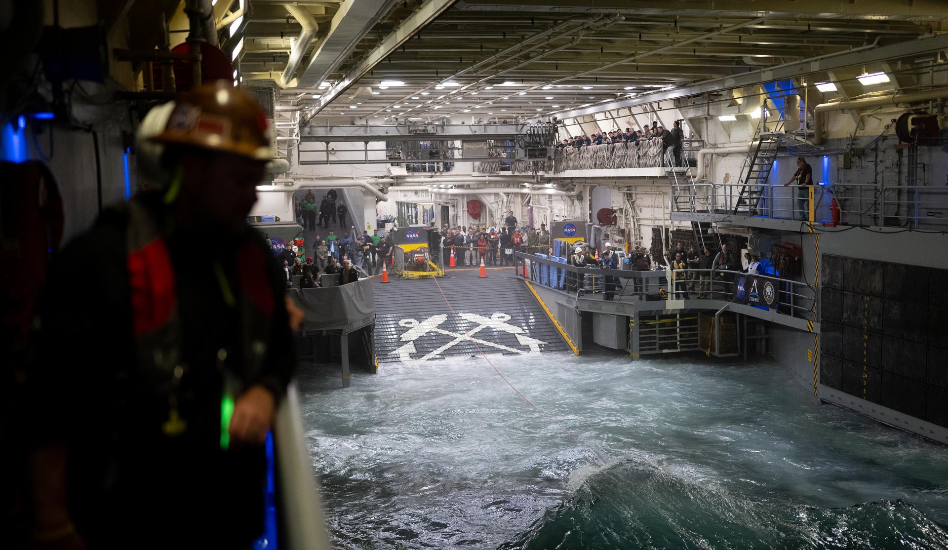 NASA and Navy personnel gather outside the safety clears in the well deck of USS John P. Murtha to watch as the agency’s Landing and Recovery team, along with U.S. Navy personnel work to recover the Orion spacecraft into the well deck of USS John P. Murtha in the Pacific Ocean off the coast of California, Friday, April 10, 2026. NASA’s Artemis II mission, which took NASA astronauts Reid Wiseman, commander; Victor Glover, pilot; Christina Koch, mission specialist; and CSA (Canadian Space Agency) astronaut Jeremy Hansen, mission specialist on a nearly 10-day journey around the Moon and back to Earth, splashed down at 5:07 p.m. PDT (8:07 p.m. EDT).  Photo Credit: (NASA/Joel Kowsky)