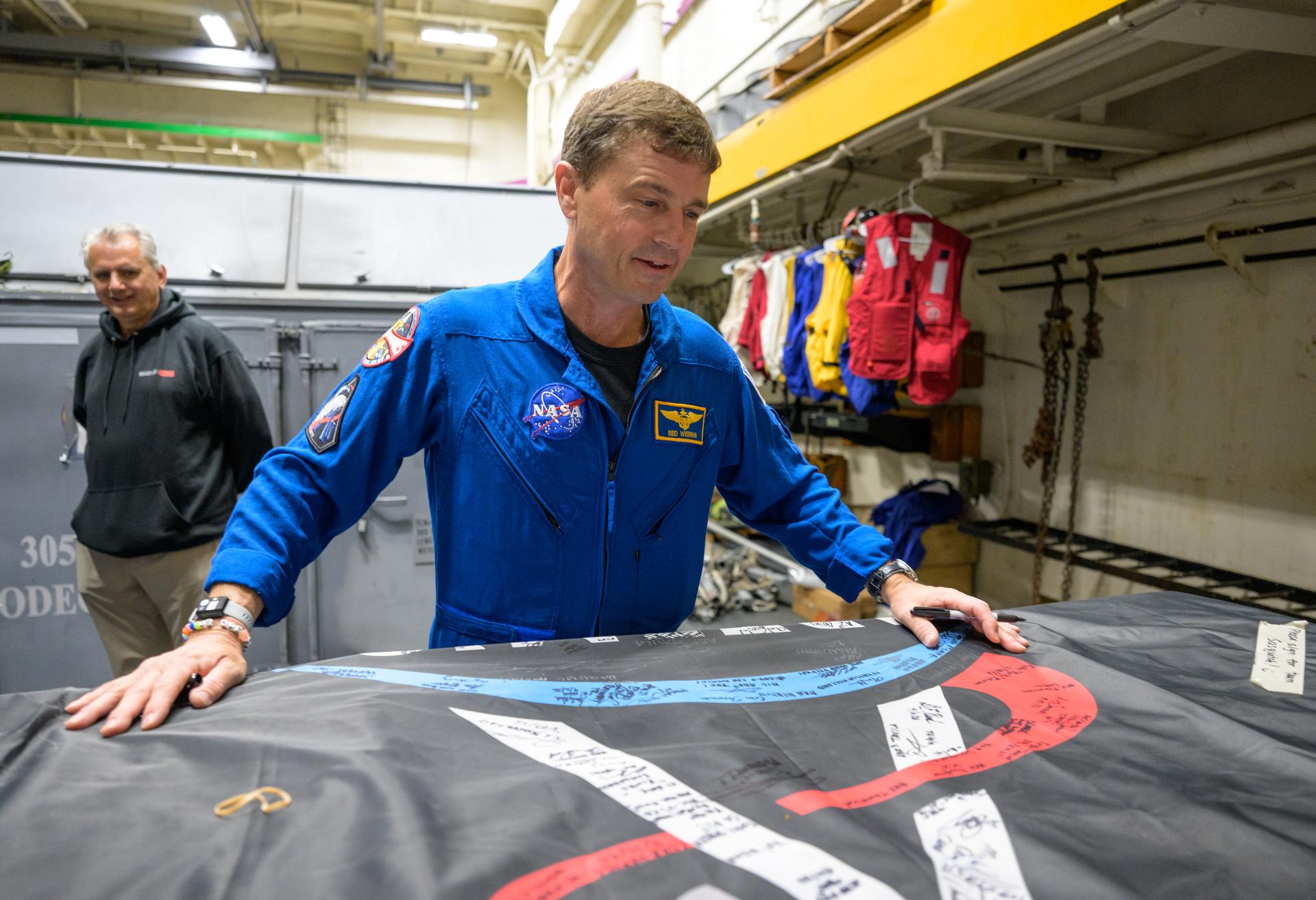 NASA astronaut Reid Wiseman, commander of the Artemis II mission looks for a place to sign an Artemis II flag onboard USS John P. Murtha, Saturday, April 11, 2026, in the Pacific Ocean off the coast of California. Wiseman, NASA astronaut Christina Koch, mission specialist; NASA astronaut Victor Glover, Artemis II pilot, and CSA (Canadian Space Agency) astronaut Jeremy Hansen, mission specialist; splashed down in the Pacific Ocean, Friday, April 10 at 5:07 p.m. PDT (8:07p.m. EDT). Photo Credit: (NASA/Bill Ingalls)