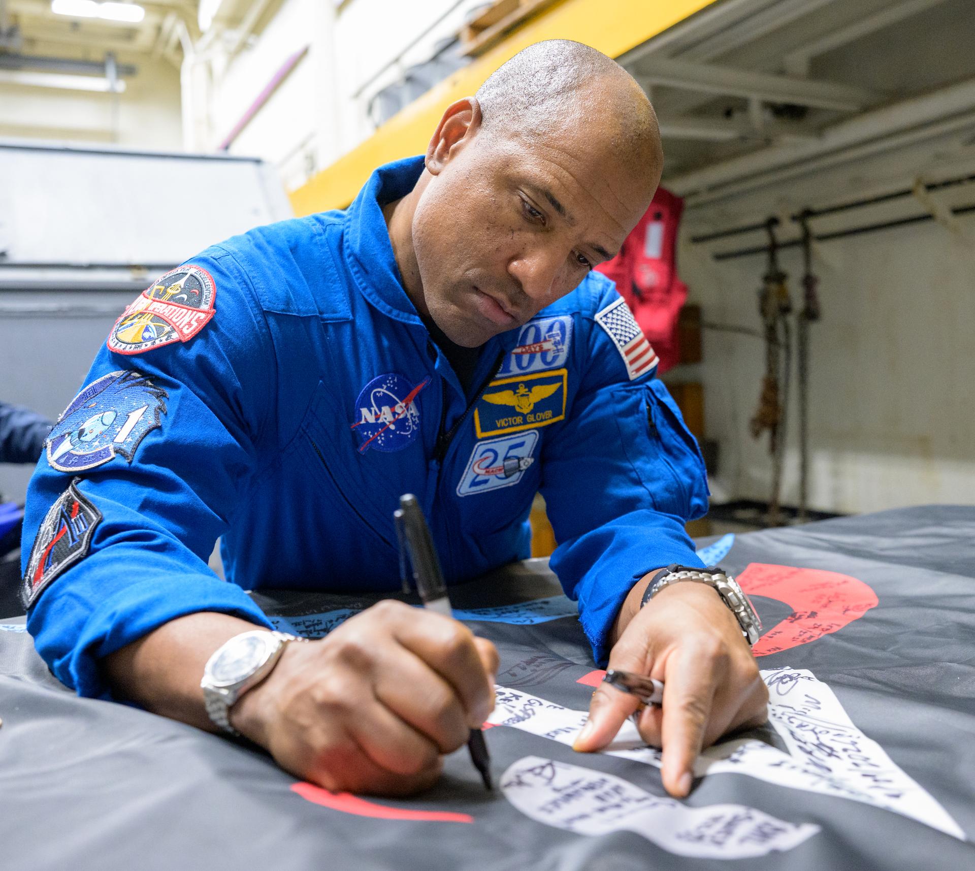 NASA astronaut Victor Glover, Artemis II pilot signs an Artemis II flag, onboard USS John P. Murtha, Saturday, April 11, 2026, in the Pacific Ocean off the coast of California. Glover, NASA astronauts Reid Wiseman, Artemis II commander, and Christina Koch, Artemis II mission specialist, along with CSA (Canadian Space Agency) astronaut Jeremy Hansen, Artemis II mission specialist, splashed down in the Pacific Ocean, Friday, April 10 at 5:07 p.m. PDT (8:07p.m. EDT). Photo Credit: (NASA/Bill Ingalls)