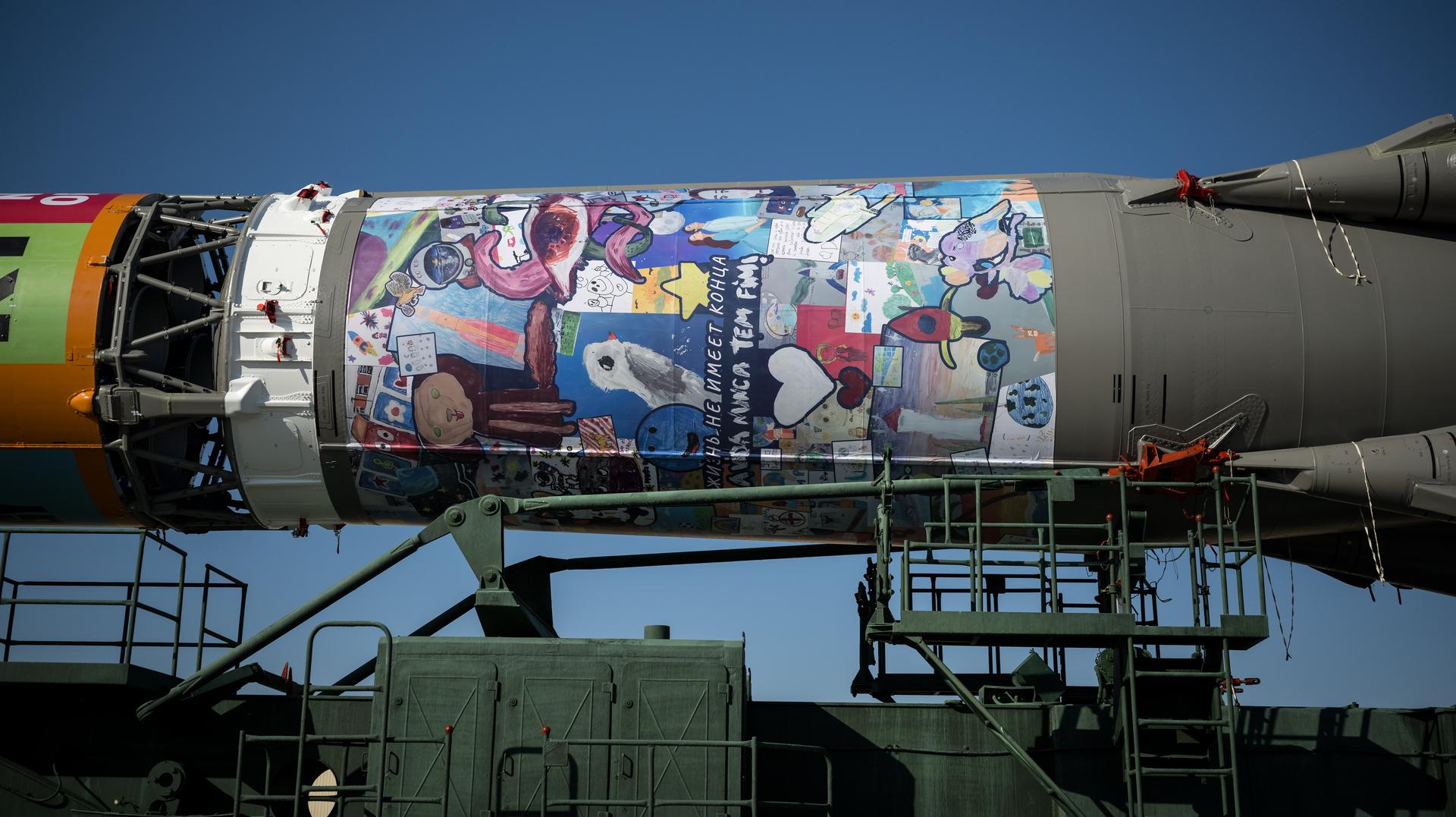 Artwork is seen on the Soyuz rocket as it is rolled out by train to the launch pad, Saturday, July 11, 2026, at Site 31 of the Baikonur Cosmodrome in Kazakhstan. Expedition 75 crewmembers: NASA astronaut Anil Menon, Roscosmos cosmonauts Pyotr Dubrov and Anna Kikina are scheduled to launch aboard their Soyuz MS-29 spacecraft on July 14. Photo Credit: (NASA/Bill Ingalls)