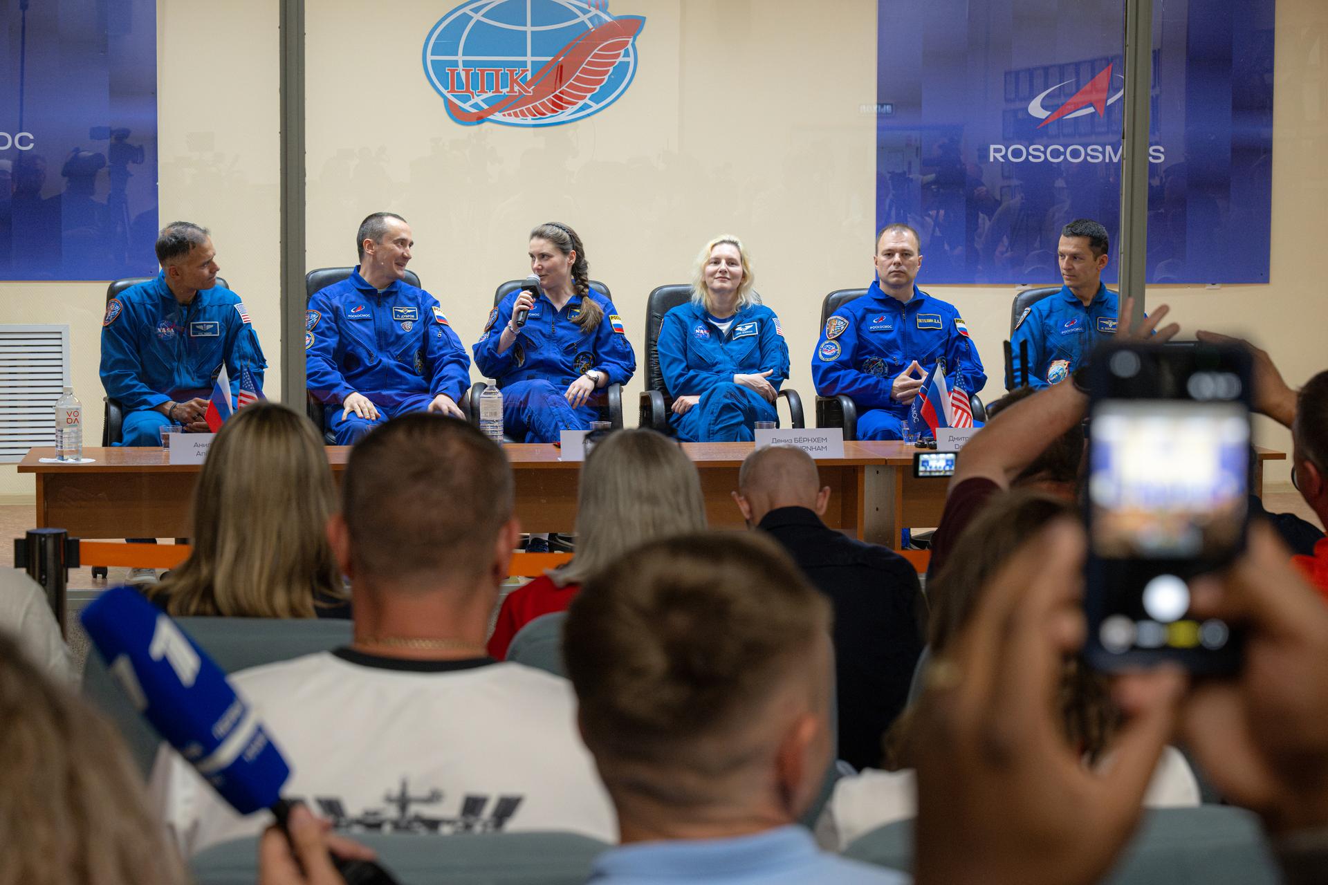 Expedition 75 crewmembers NASA astronaut Anil Menon, left, Roscosmos cosmonauts Pyotr Dubrov and Anna Kikina, along with backup crew members Deniz Burnham of NASA and Dmitry Petelin and Konstantin Borisov of Roscosmos, right, are seen in quarantine, behind glass, during a press conference, Monday, July 13, 2026, at the Cosmonaut Hotel in Baikonur, Kazakhstan. Menon, Dubrov, and Kikina are scheduled to launch aboard the Soyuz MS-29 spacecraft to the International Space Station on July 14. Photo Credit: (NASA/John Kraus)