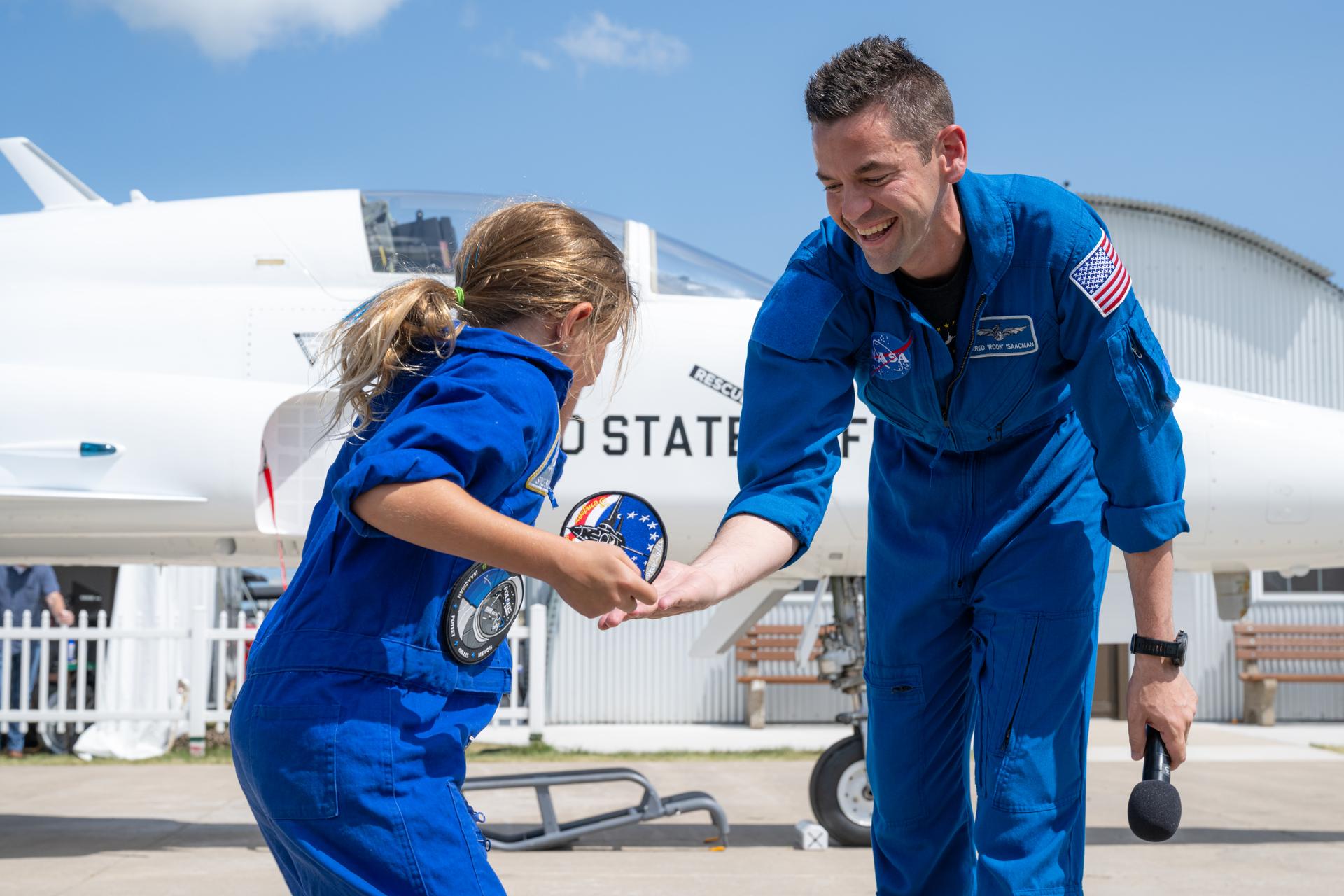 NASA Administrator Jared Isaacman gives a patch from his flight suit and offers a high five to a young aviation enthusiast during a “Warbirds in Review” presentation, Thursday, July 23, 2026, at EAA AirVenture Oshkosh at Wittman Regional Airport in Oshkosh, Wis. Isaacman discussed NASA’s efforts in air and space, his aviation experience, and more. Photo Credit: (NASA/John Kraus)