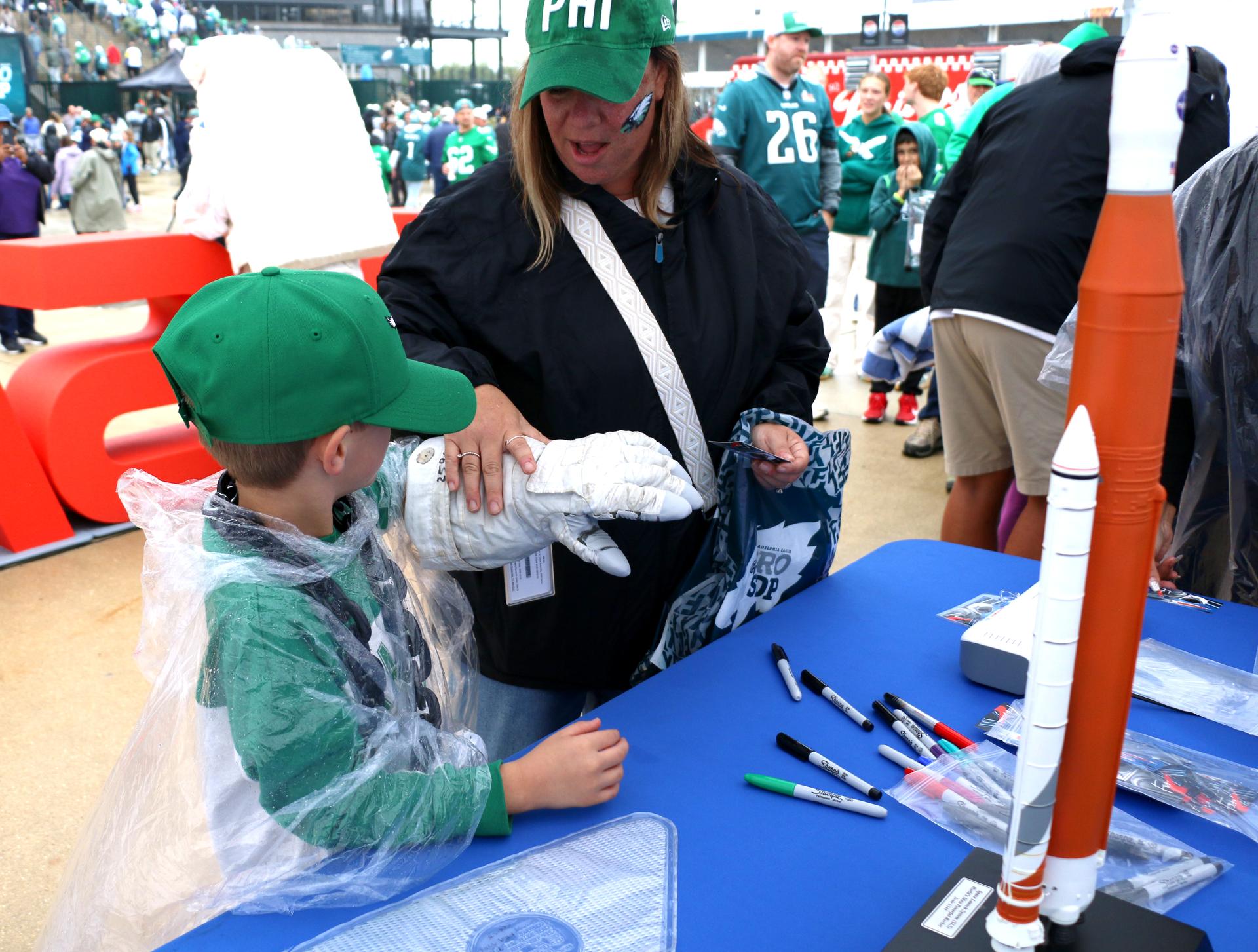 Two fans try on an astronaut glove at the NASA activation at the NFL game between the Philadelphia Eagles and the Los Angeles Rams at Lincoln Financial Field on Sunday, Oct. 4, 2026, in Philadelphia. NASA’s engagement at NFL games is part of the agency's Inspiration Tour, aimed at strengthening connections between NASA and its partners and showcasing innovation in air and space in the lead up to the MAX POWER aerospace technology expo and airshow at the agency's Kennedy Space Center in early November. Photo Credit: (NASA/Thalia Patrinos)