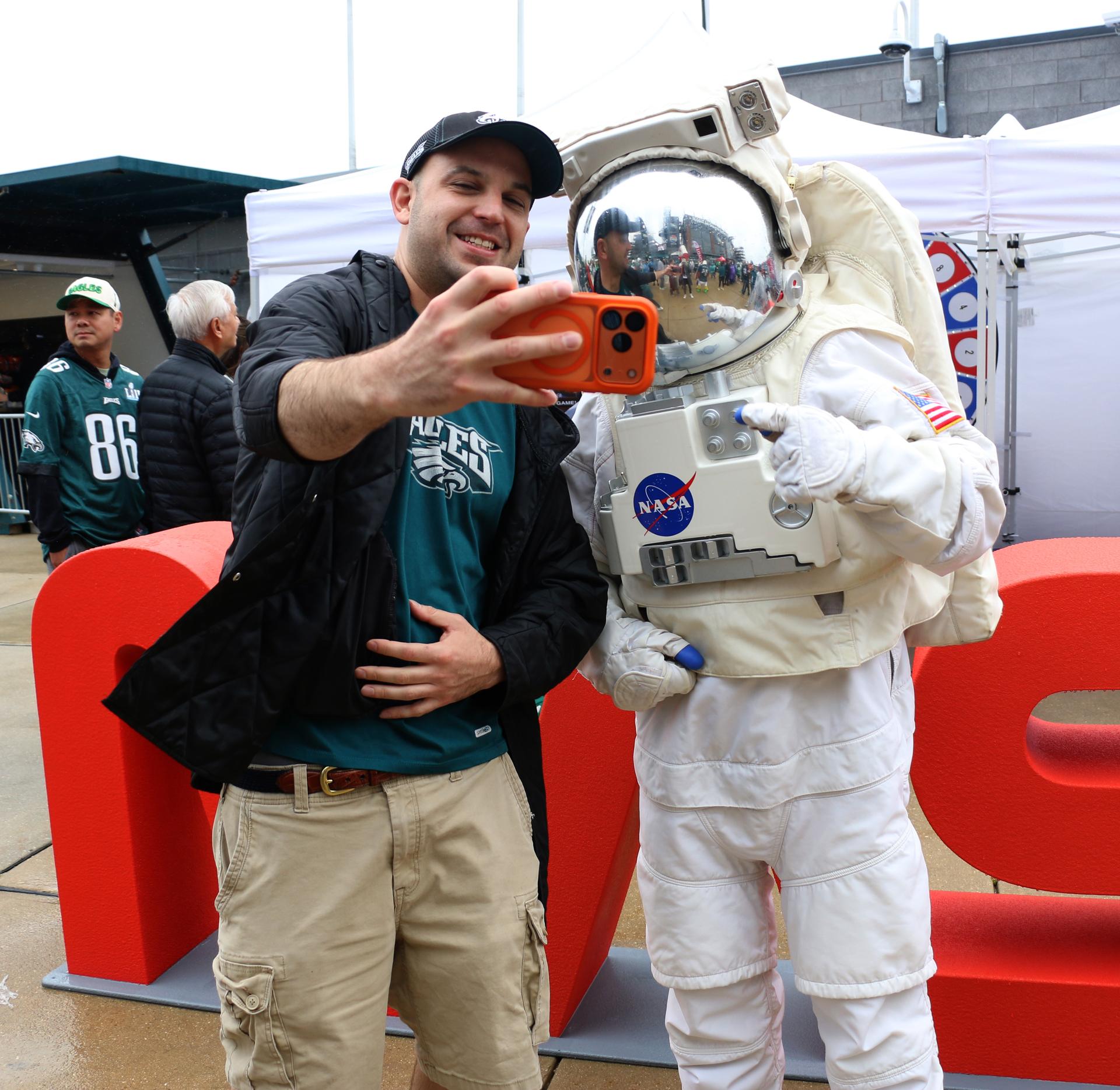 A fan takes a selfie with the astronaut at the NASA activation at the NFL game between the Philadelphia Eagles and the Los Angeles Rams at Lincoln Financial Field on Sunday, Oct. 4, 2026, in Philadelphia. NASA’s engagement at NFL games is part of the agency's Inspiration Tour, aimed at strengthening connections between NASA and its partners and showcasing innovation in air and space in the lead up to the MAX POWER aerospace technology expo and airshow at the agency's Kennedy Space Center in early November. Photo Credit: (NASA/Thalia Patrinos)
