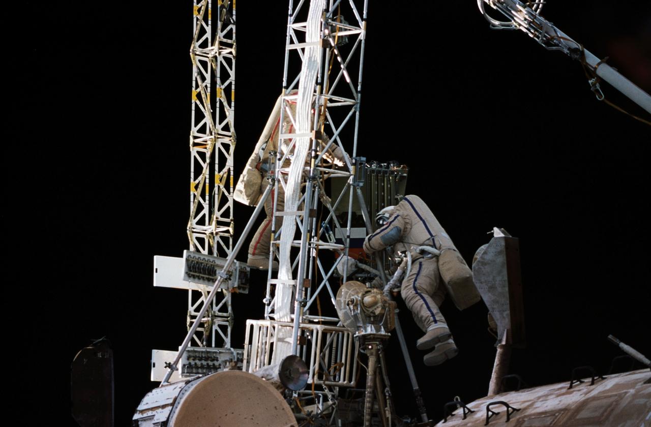 NM21-382-010 (For Release October 1996) --- Mir 21 commander Yury I. Onufrienko (left), wearing a red stripe on his Russian Orlan spacesuit, and Mir 21 flight engineer Yury V. Usachev (blue stripe on Orlan)traverse an existing truss on the Kvant module with a folded truss in tow.