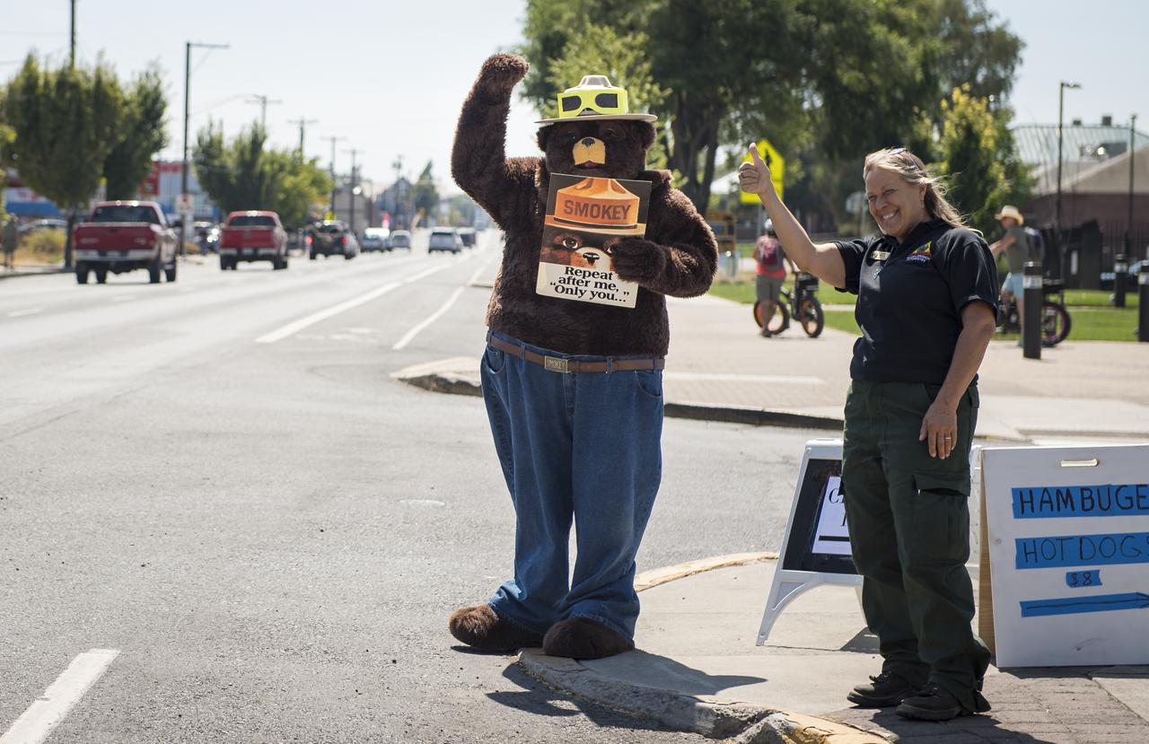 Smokey Bear, left, and Judy Reese of the Crooked River National Grassland, right, wave at passersby as crowds arrive to watch the total solar eclipse in Madras, Oregon on Saturday, August 19, 2017. The eclipse will be sweeping across a narrow portion of the contiguous United States from Lincoln Beach, Oregon to Charleston, South Carolina on August 21. A partial solar eclipse will be visible across the entire North American continent along with parts of South America, Africa, and Europe.  Photo Credit: (NASA/Aubrey Gemignani)