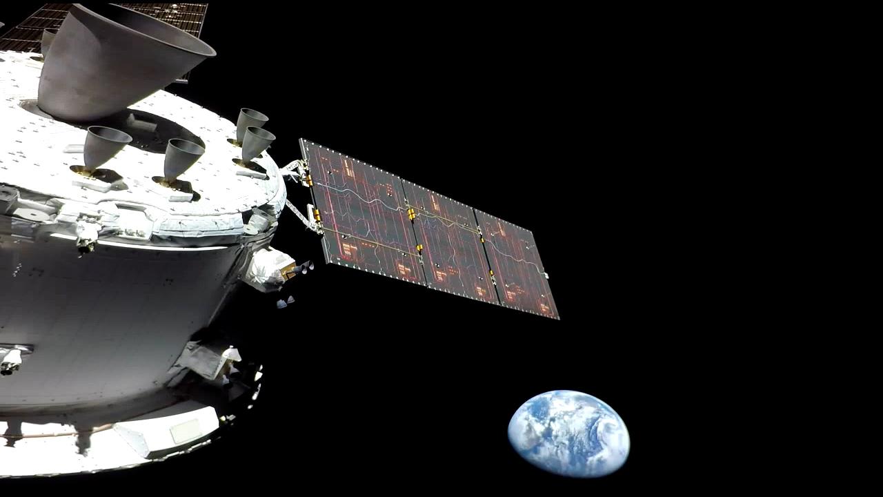 The white and metallic grey Orion spacecraft with solar arrays deployed looks back at Earth in the distance. The blue and white swirls of Earth’s surface stand in stark contrast to the blackness of space.