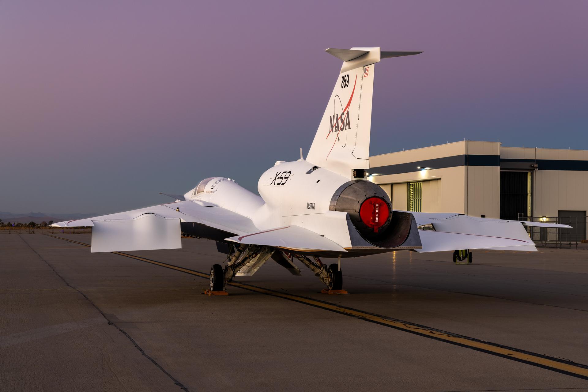 NASA’s X-59 sits on the ramp of Lockheed Martin’s Skunk Works facility, facing toward the site’s main hangar during sunset.