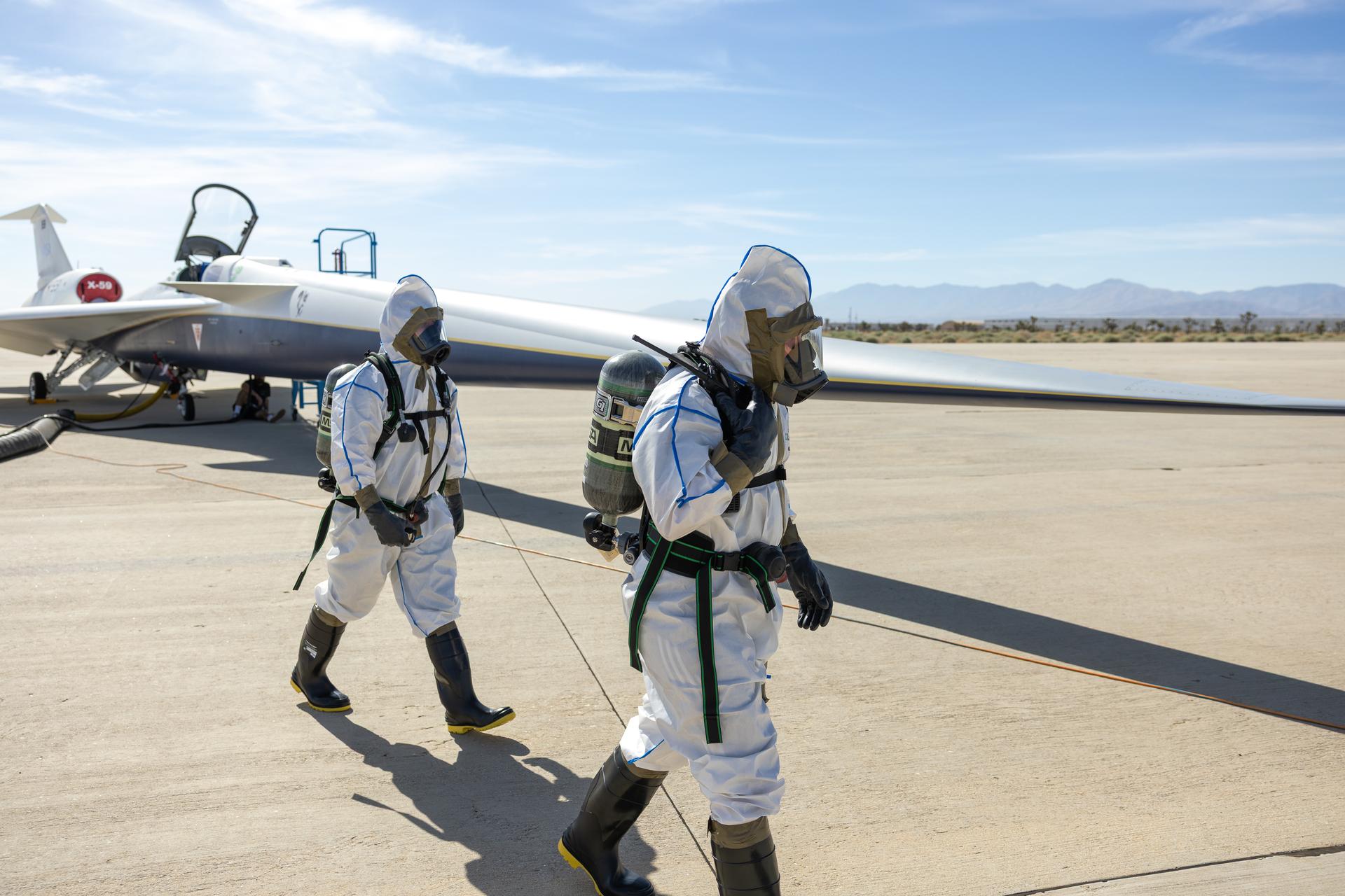 Two maintainers in white protective suits, air tanks, masks, rubber gloves, and boots walk around the front of NASA’s X-59 quiet supersonic research aircraft during a hydrazine safety check at U.S. Air Force Plant 42 in Palmdale, California, on Aug. 18, 2025. The sky is mostly clear, with some vegetation and mountains visible in the background. The aircraft has its canopy open, a ladder positioned on the right side of its fuselage, and air ducts extending from the aircraft.