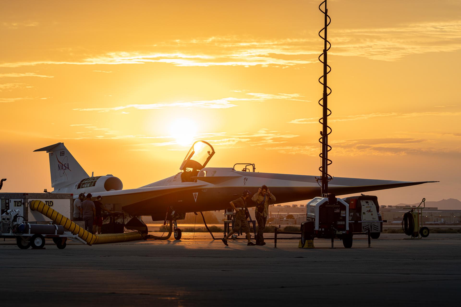 NASA’s X-59 quiet supersonic research aircraft is seen on the ramp at sunrise with its canopy open. Orange and gold light filters through the clouds behind the aircraft, highlighting the aircraft’s shape. Several ground crew members are visible around the aircraft, along with support carts. A vertical light pole with a coiled cable can be seen in the foreground.