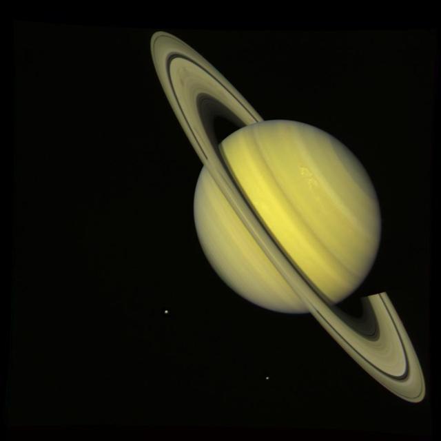 NASA Voyager 2 took this true color photograph of Saturn on July 21, 1981. The moons Rhea and Dione appear as blue dots to the south and southeast of Saturn, respectively.