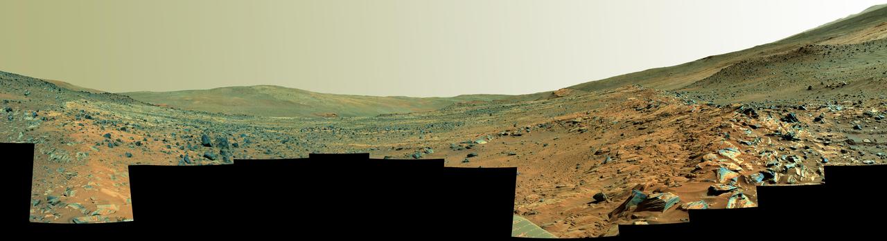 This panorama shows two rock-strewn slopes on the left and right sides of a broad, U-shaped dip in the middle. The sandy surface in front of the rover is reddish brown; individual rocks and more distant features are blue-gray