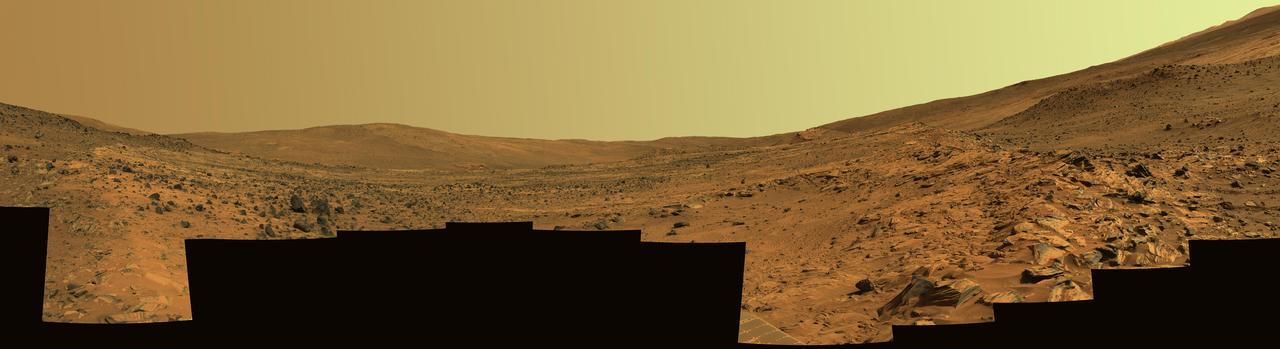 This panorama shows two reddish-brown, rock-strewn slopes on the left and right sides of a broad, U-shaped dip in the middle. In the distance are the broad slopes of McCool Hill. Above that is an orangish-yellow sky.