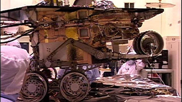 This image taken at NASA Jet Propulsion Laboratory shows engineers testing the rover stand-up motions. The rover is in an elevated pose, preparing to fold down its wheels, descend and stand up.