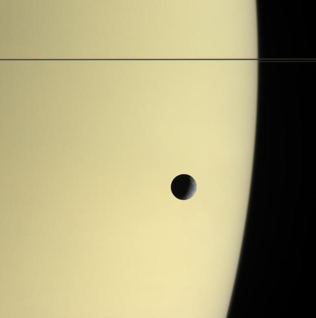 Tethys floats before the massive, golden-hued globe of Saturn in this natural color view. The thin, dark line of the rings curves around the horizon at top