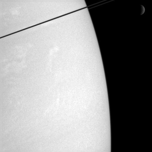 Whiffs of cloud dance in Saturn atmosphere, while the dim crescent of  Rhea 1,528 kilometers, or 949 miles across hangs in the distance. The dark ringplane cuts a diagonal across the top left corner of this view