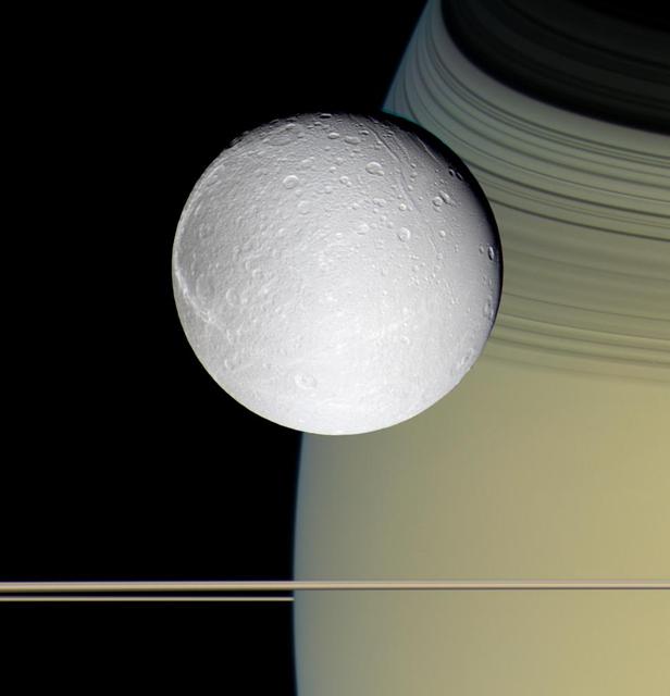 Speeding toward pale, icy Dione, Cassini view is enriched by the tranquil gold and blue hues of Saturn in the distance. The horizontal stripes near the bottom of the image are Saturn rings.