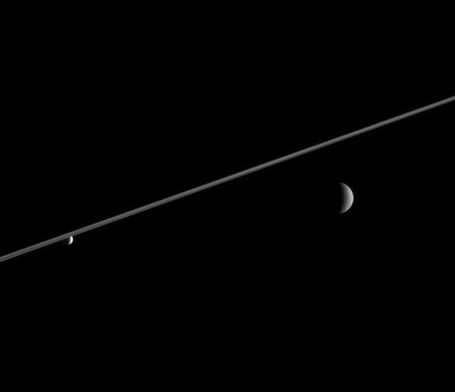 Rhea and Enceladus hover in the distance beyond Saturn ringplane. Enceladus left, bathed in icy particles from Saturn E ring, appears noticeably brighter than Rhea