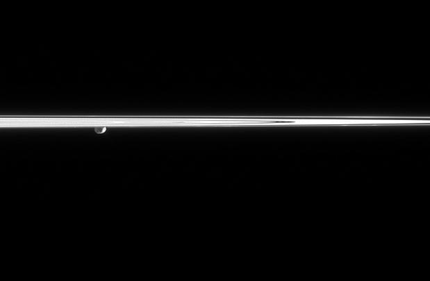 Janus peeks out from beneath the ringplane, partially lit here by  reflected light from Saturn. A couple of craters can be seen on the moon  surface. To the right, two faint clumps of material can be seen in the  dynamic F ring