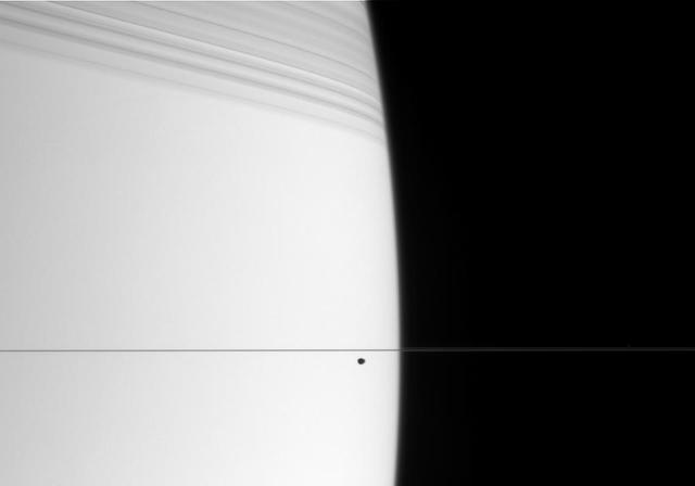 The small, dark form of Janus cruises along in front of bright Saturn. The edge-on rings cast dramatic shadows onto the northern hemisphere