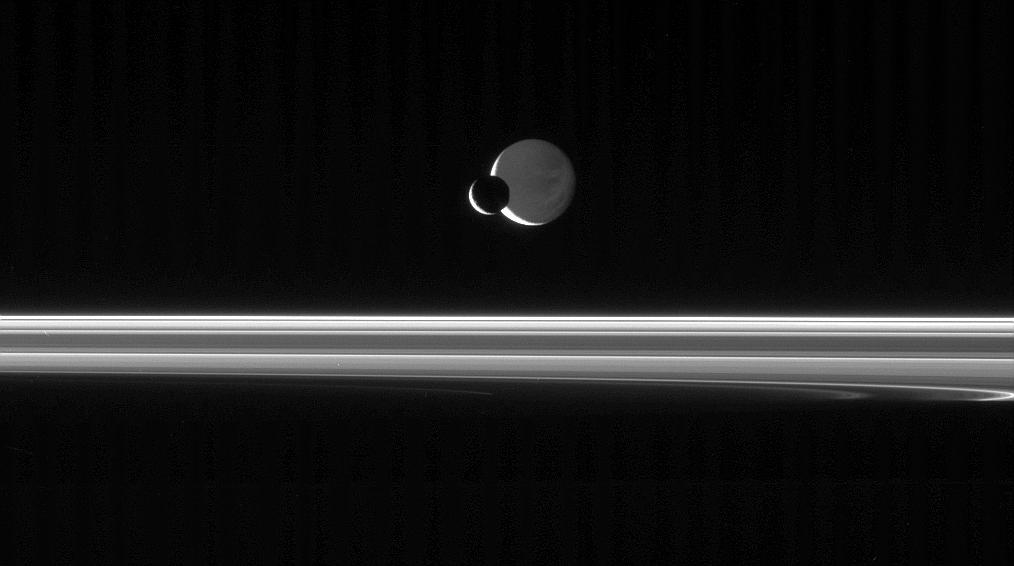 The Cassini spacecraft looks across the unlit ringplane as Mimas glides  silently in front of Dione
