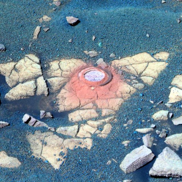 This false-color image shows a circular indentation in a flat-topped rock surface. Around the edge of the hole is a fine layer of reddish dust. The rock is light tan and has a moderately cracked the surface. Around it is a layer of bluish sand and pebbles