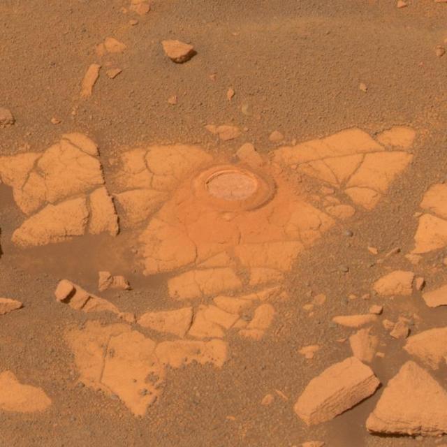 This true-color image shows a circular indentation in a flat-topped rock surface. Around the edge of the hole is a fine layer of dust. The rock has a moderately cracked the surface. Around it is a layer of sand and pebbles. The view is reddish brown